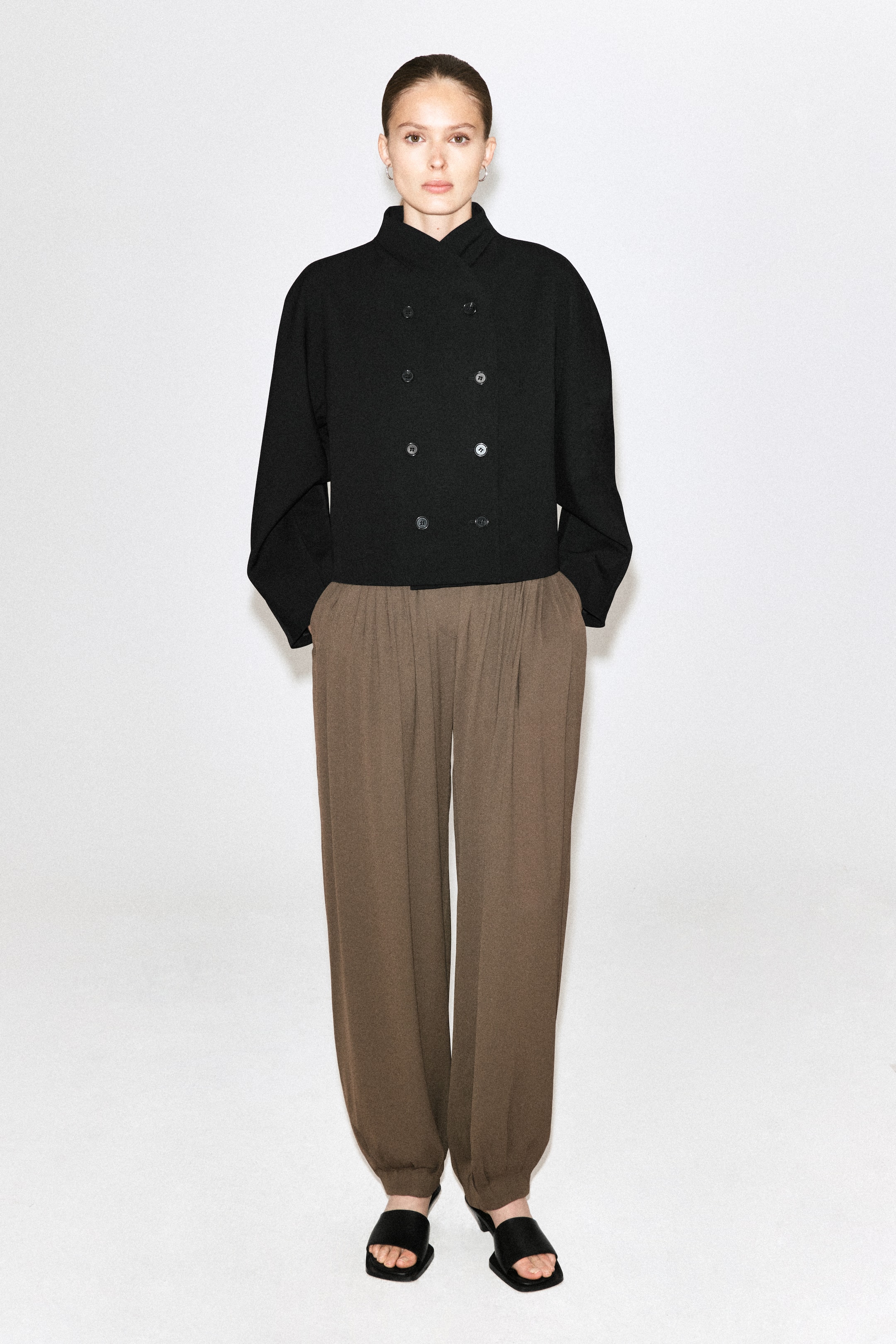 Agrandir l'image: Le mannequin porte un pantalon ballon plissé en crêpe marron, à la silhouette décontractée avec des plis à la taille et des chevilles froncées. Le pantalon est associé à une veste noire croisée et à des sandales noires ouvertes à l’avant type mules.