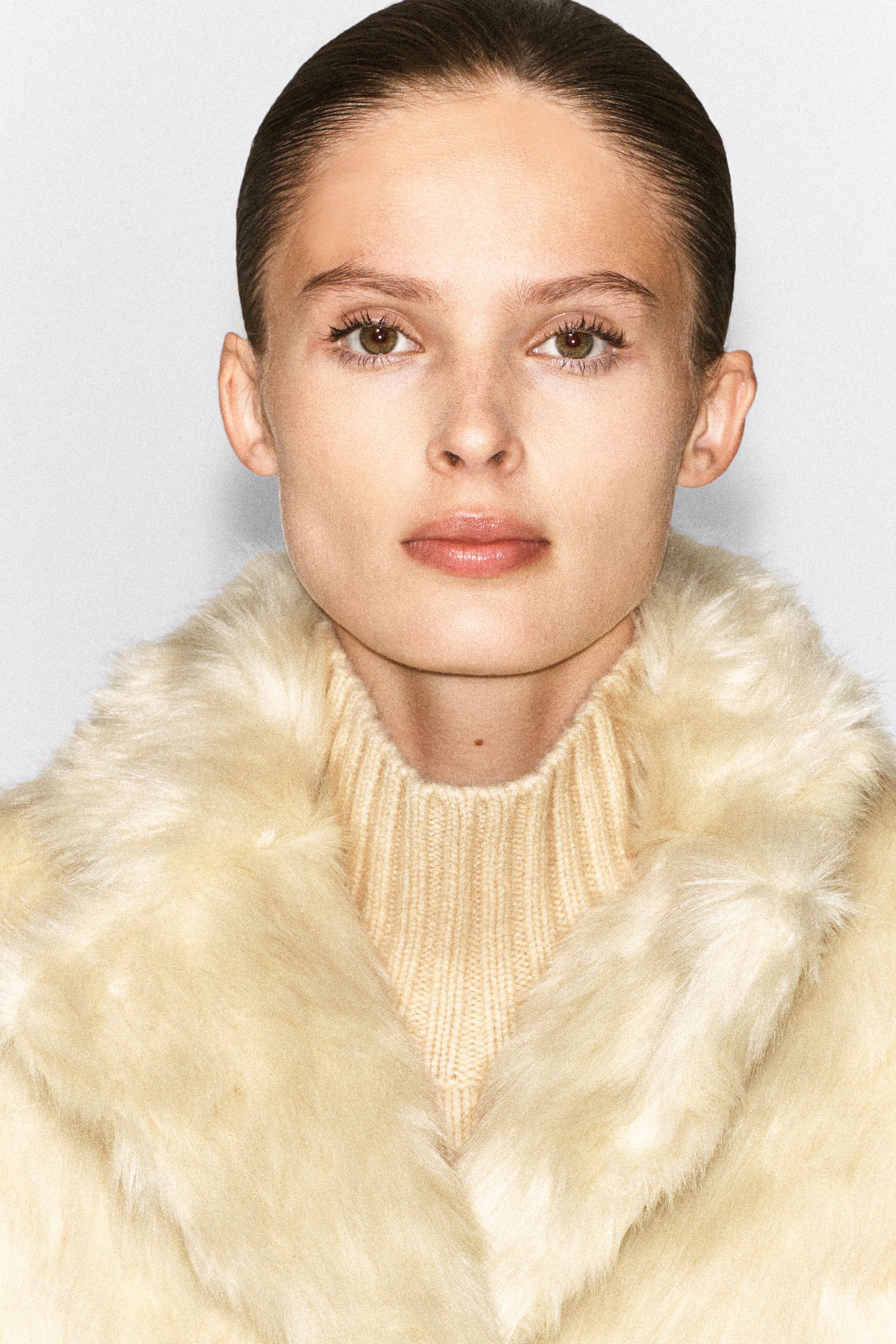 The model is wearing a cream-colored, faux-fur jacket with a wide collar, layered over a beige ribbed turtleneck sweater.