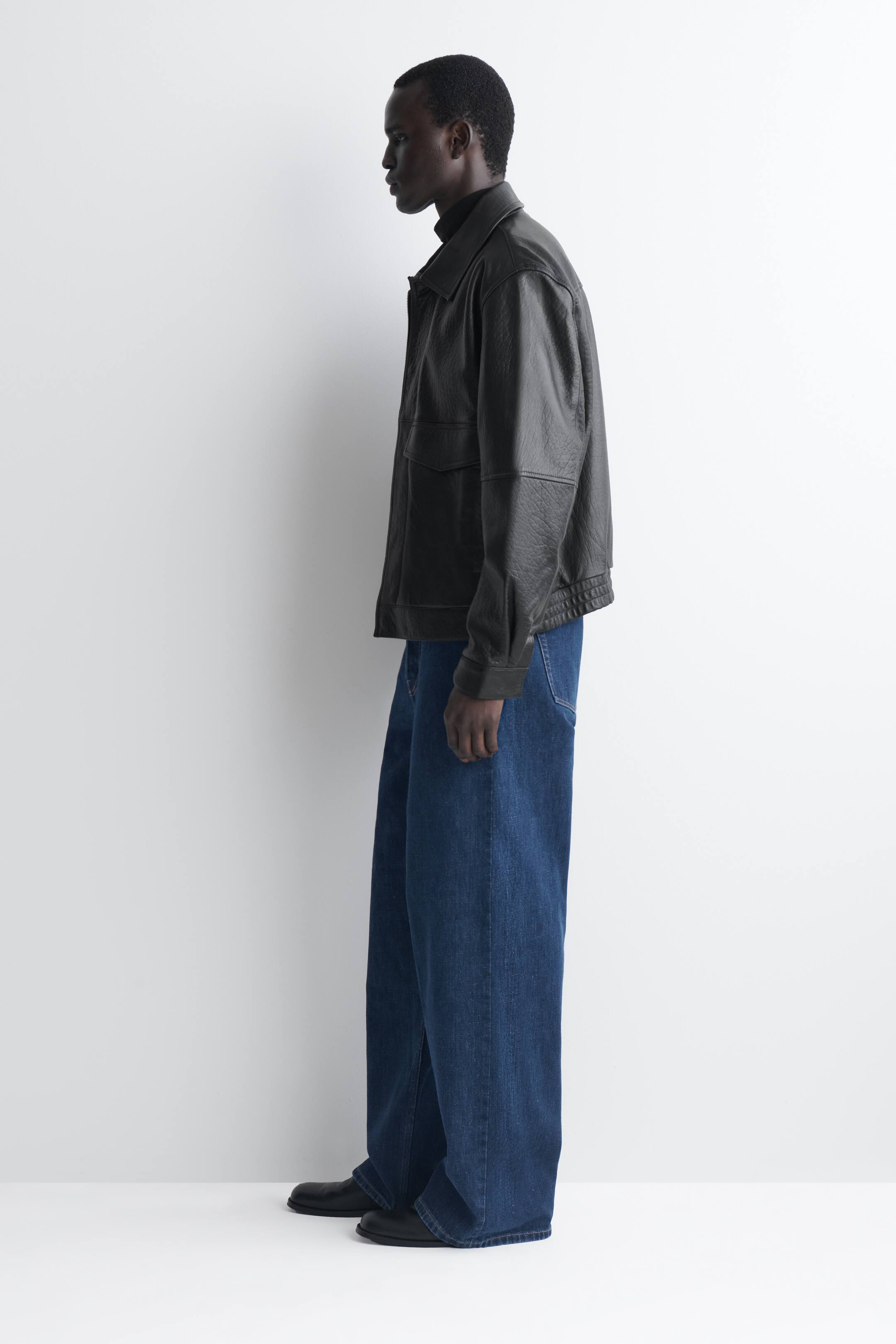 Visualizza l'immagine ingrandita: Questi jeans in denim blu a gamba larga cadono con una vestibilità ampia e lunga, appoggiandosi leggermente sulle scarpe nere mentre il modello è in piedi rivolto verso sinistra per mettere in risalto la loro silhouette morbida. I jeans a vita media sono abbinati a una giacca in pelle nera e a un dolcevita nero.