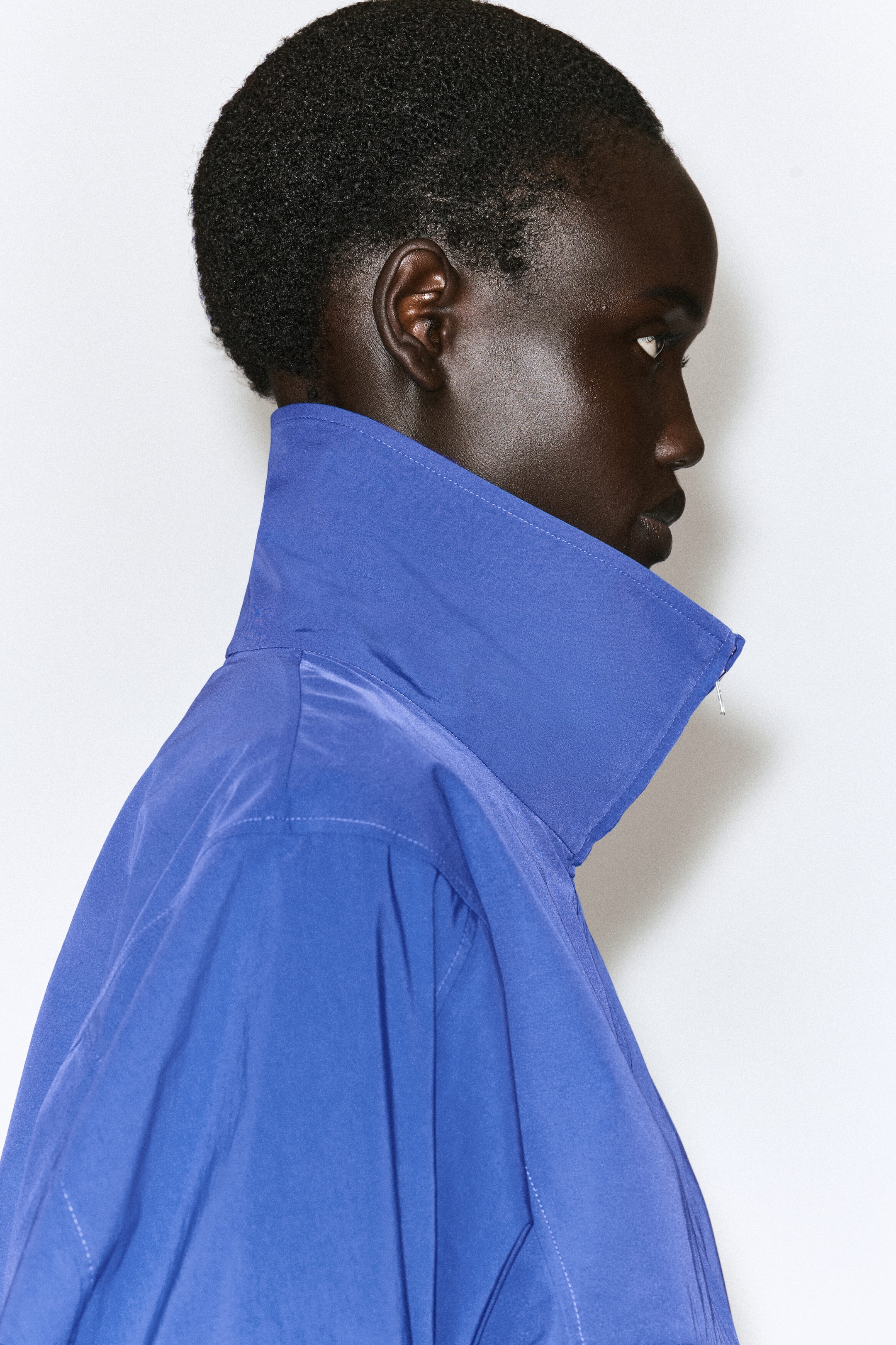A blue zip-up shirt featuring a high, structured stand collar and topstitched seams is shown in profile, facing right. A blue zip-up shirt featuring a high, structured stand collar and topstitched seams is shown in profile, facing right.