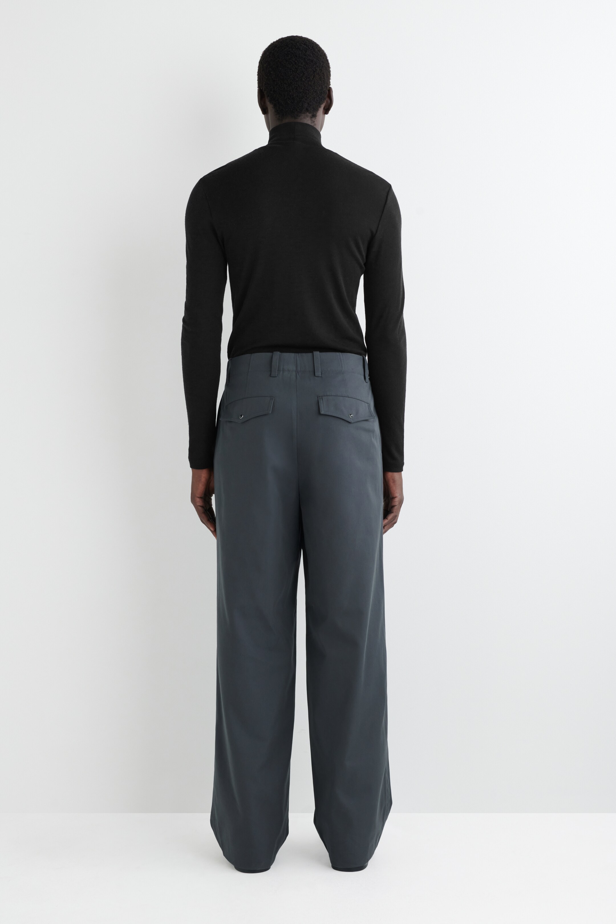 View larger image: A black long-sleeved turtleneck shirt is being worn, seen from the back, tucked into gray wide-leg trousers. The trousers are featuring two buttoned flap pockets on the rear and belt loops, showing a loose, flowing fit.