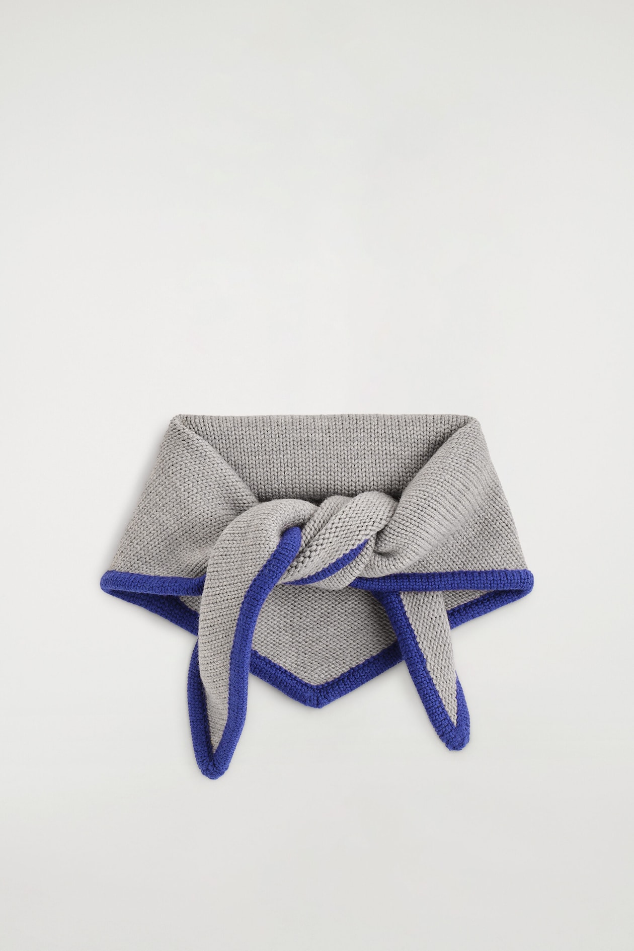 The contrast-tipped merino wool triangle scarf is shown from a front-facing angle, displaying a light grey knitted texture with a vibrant blue contrast border. The scarf is resting in a loose knot at the centre, with its triangular tip pointing downwards beneath the two hanging ends.