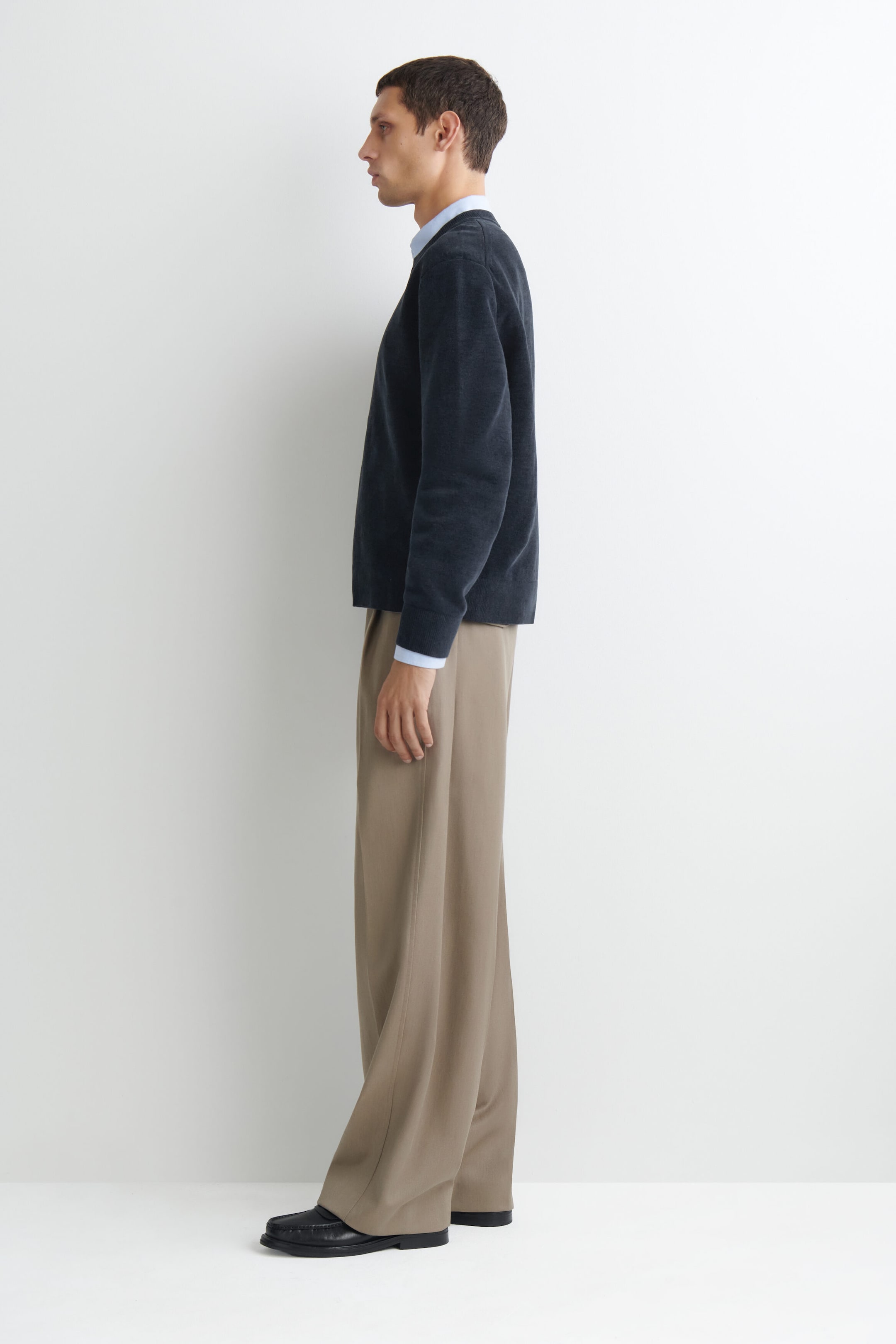 A model facing left is wearing a dark gray cotton-chenille crewneck jumper, highlighting its soft, textured finish, ribbed cuffs, and relaxed fit. The jumper is layered over a light blue collared shirt, with the collar and cuffs visible, and is paired with wide-leg beige trousers. A model facing left is wearing a dark gray cotton-chenille crewneck jumper, highlighting its soft, textured finish, ribbed cuffs, and relaxed fit. The jumper is layered over a light blue collared shirt, with the collar and cuffs visible, and is paired with wide-leg beige trousers.