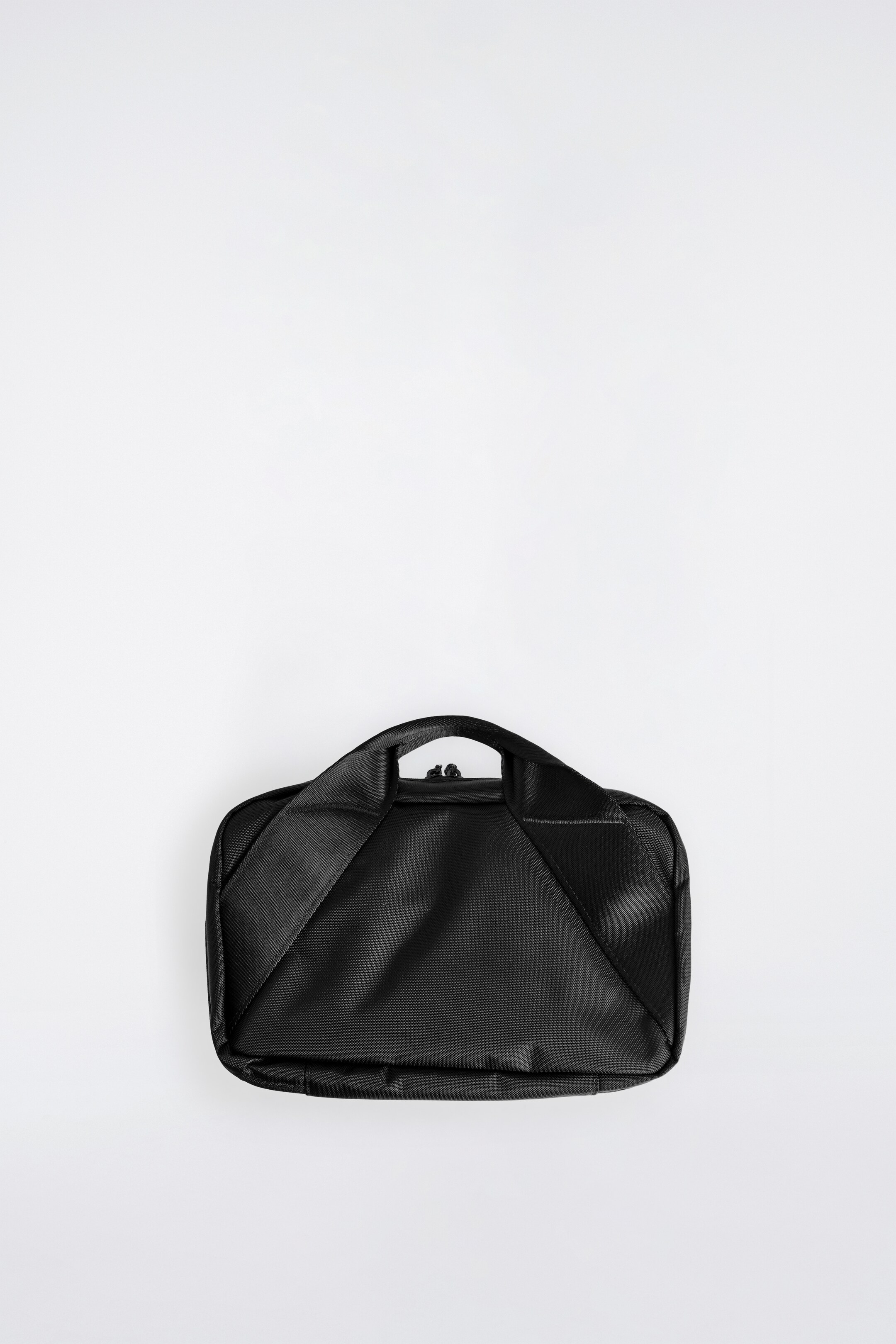 Agrandir l'image: Une trousse de toilette noire est présentée de son côté inférieur, à plat. Une poignée supérieure, formée de matériau de sangle noir, s'étend en diagonale sur sa surface arrière, et une fermeture éclair est visible le long du bord supérieur.