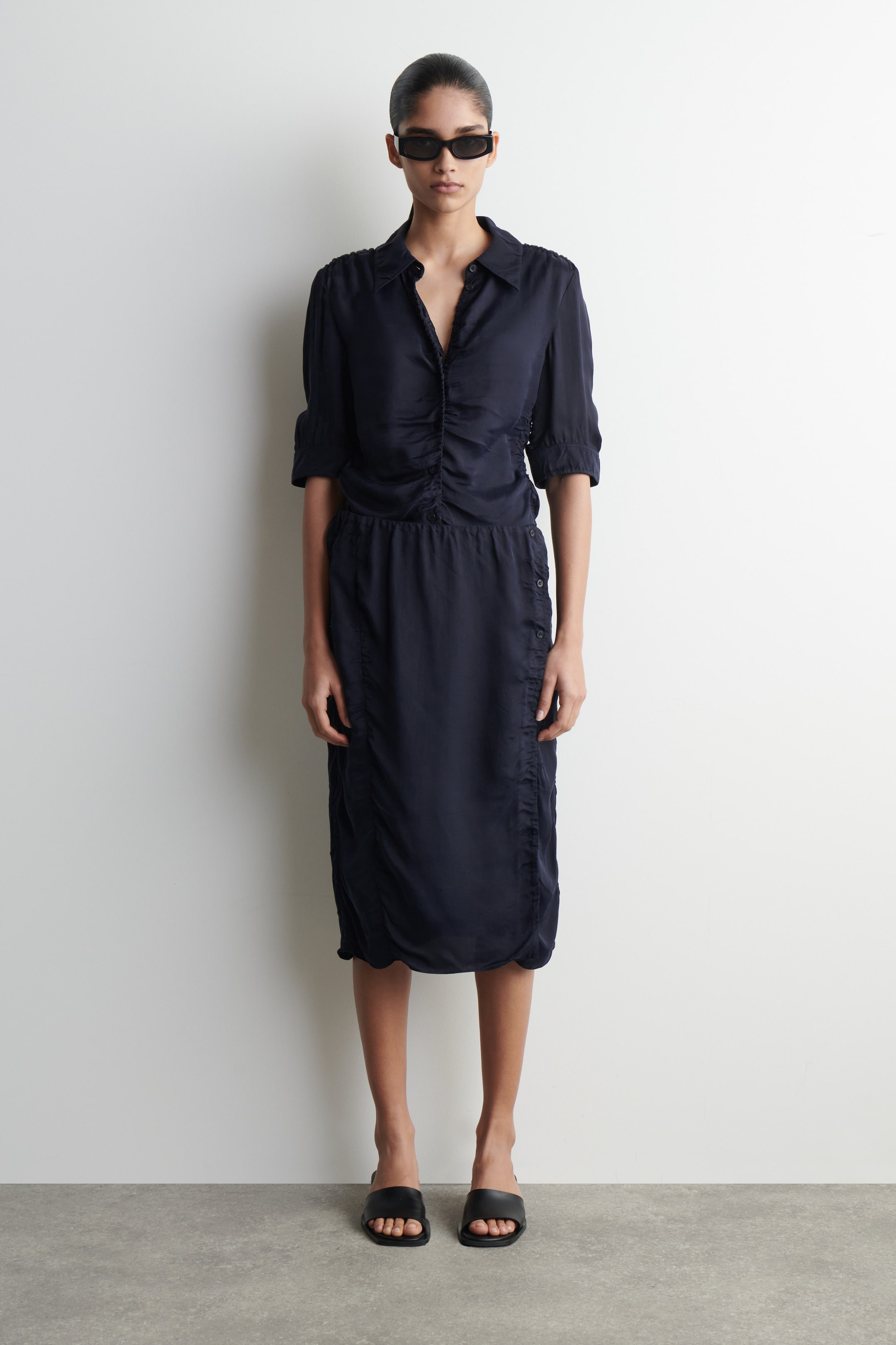 View larger image: A dark navy midi dress featuring a shirt collar, an open V-neckline, and elbow-length sleeves with gathered cuffs. The bodice showcases front ruching, and the skirt is designed with side button plackets, additional side ruching, and a gathered hemline.