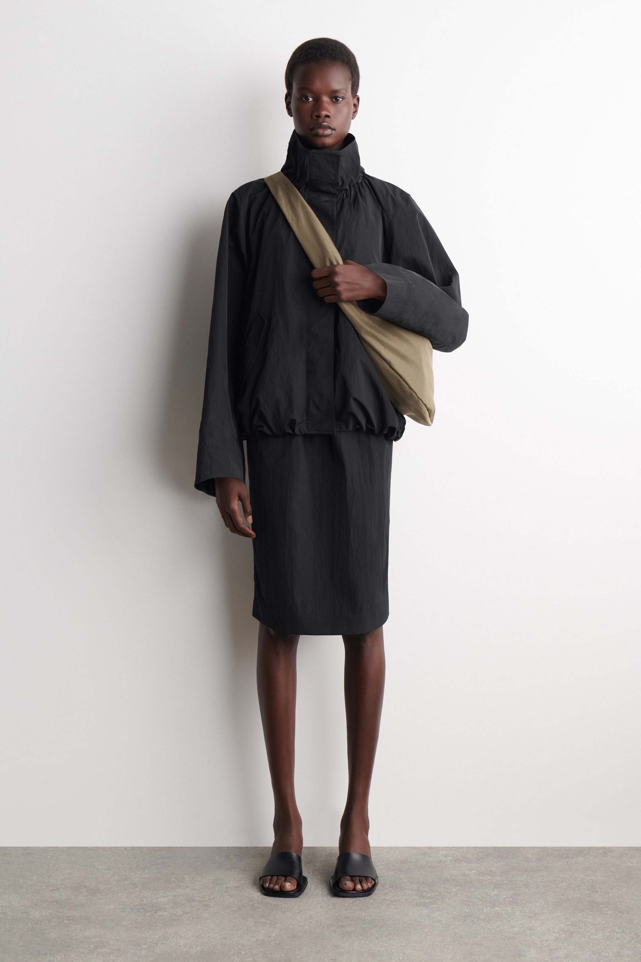 Visa större bild: En svart löst sittande blousonjacka med en hög rynkad ståkrage och en elastisk fåll bärs över en svart knälång kjol. En mjuk, ljusbeige crossbody-väska hänger över jackan, och svarta fyrkantiga tofflor med öppen tå bärs, alla vända framåt.