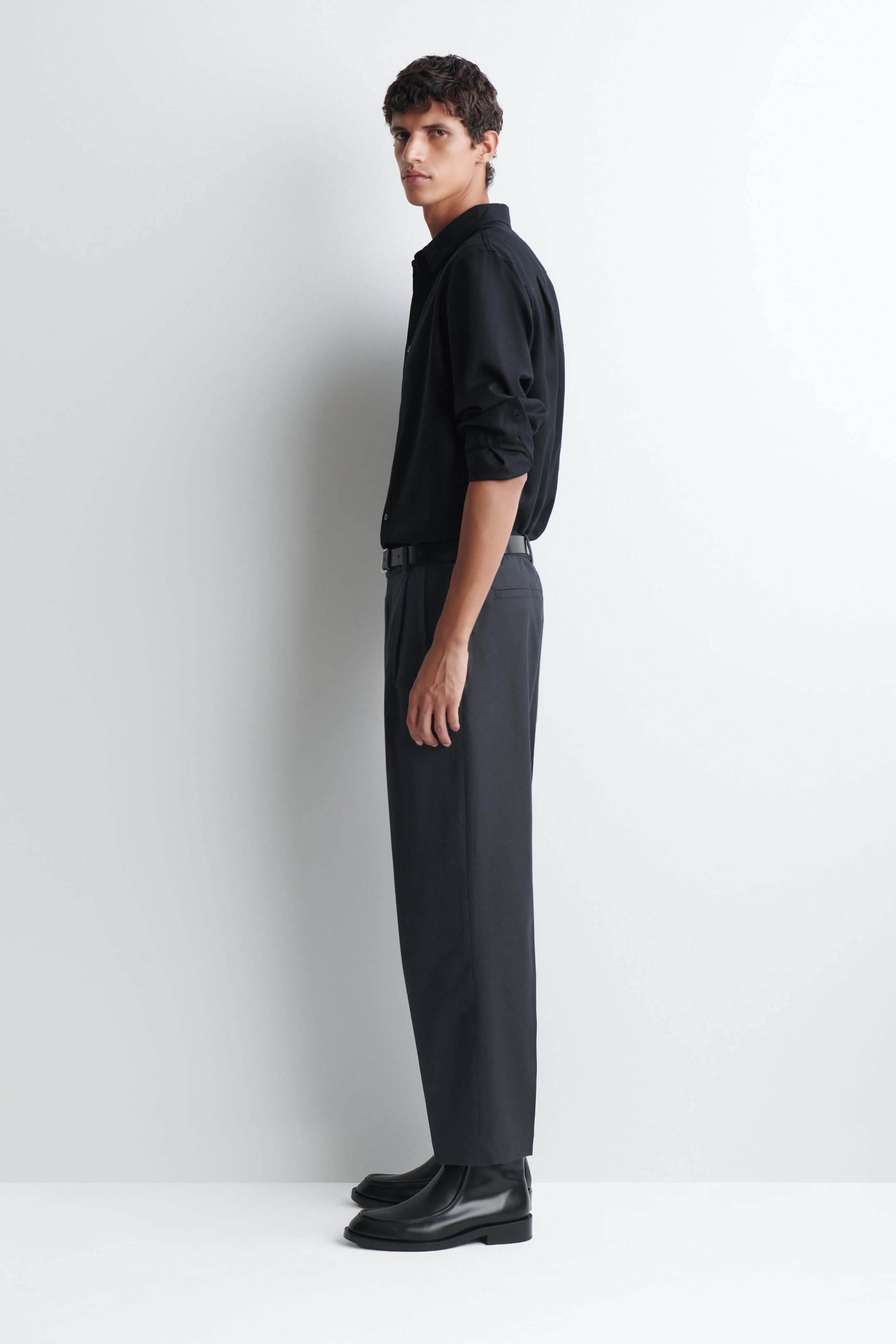 The model is wearing dark gray pleated trousers featuring a straight-leg silhouette and a high-rise waist. The trousers are styled with a tucked-in black button-up shirt, a black leather belt, and black boots. The model is wearing dark gray pleated trousers featuring a straight-leg silhouette and a high-rise waist. The trousers are styled with a tucked-in black button-up shirt, a black leather belt, and black boots.