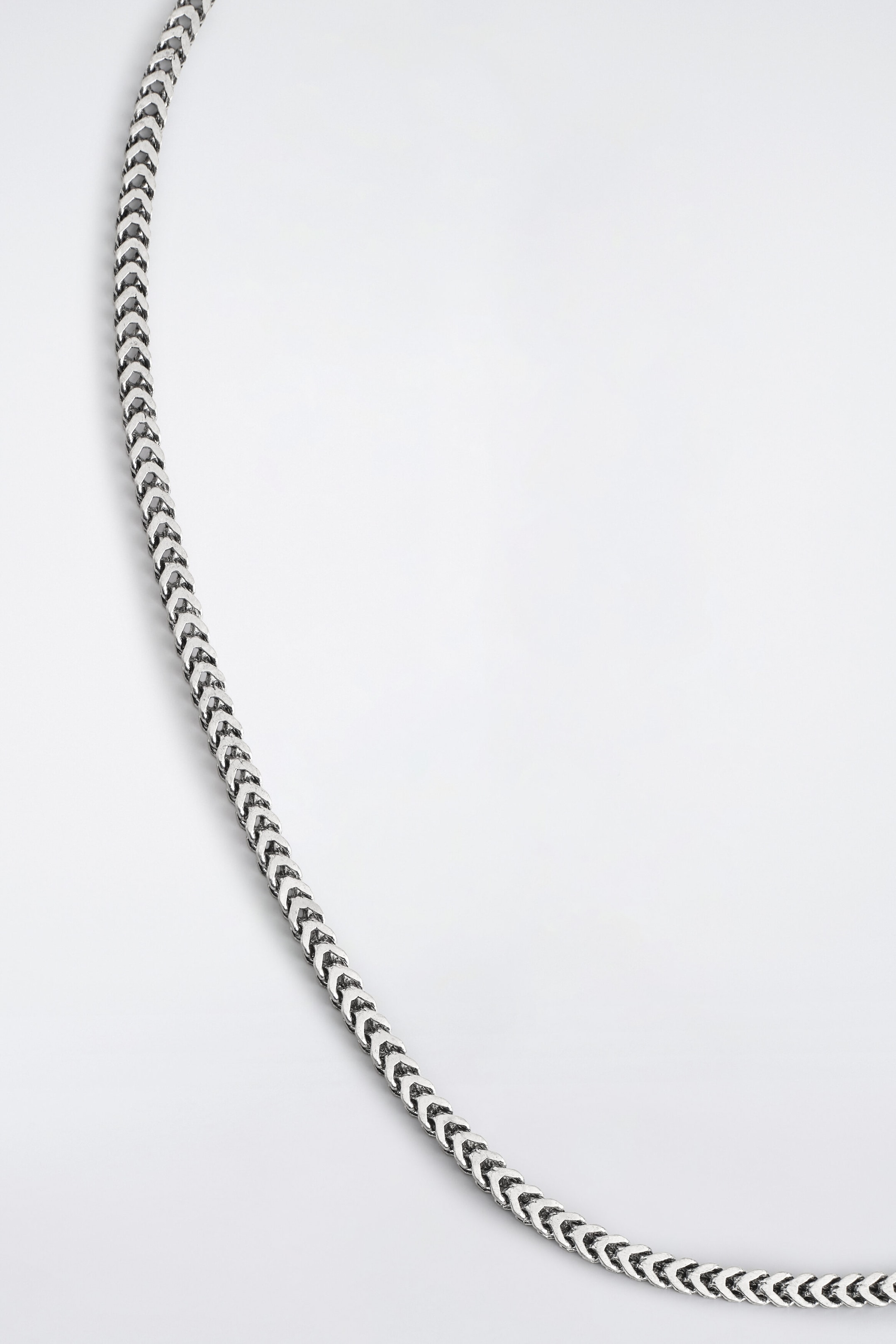 A silver-tone metal chain featuring interlocking, chevron-patterned links curves from the upper left to the lower right. The chain has a polished finish with darkened, recessed details that accentuate its textured, three-dimensional structure.