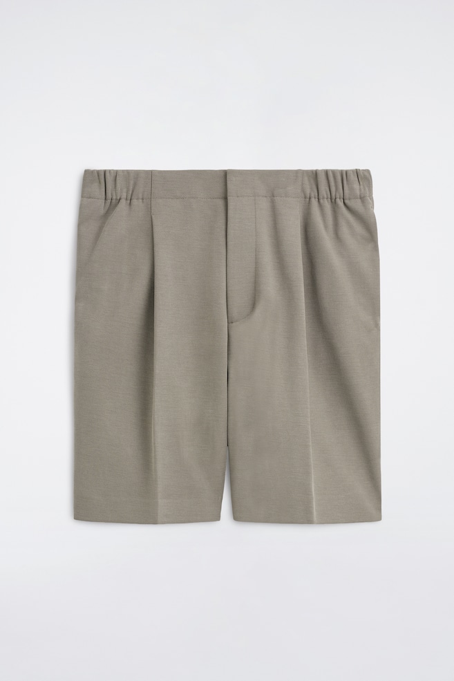 Le short Tailored Bonded-Twill est présenté de face, mettant en valeur un tissu taupe-gris structuré avec une ceinture plate sur le devant et des empiècements latéraux élastiqués. Ce short présente des plis simples qui se prolongent en plis marqués au centre le long des jambes, ainsi que des poches latérales et une braguette frontale dissimulée. - 1