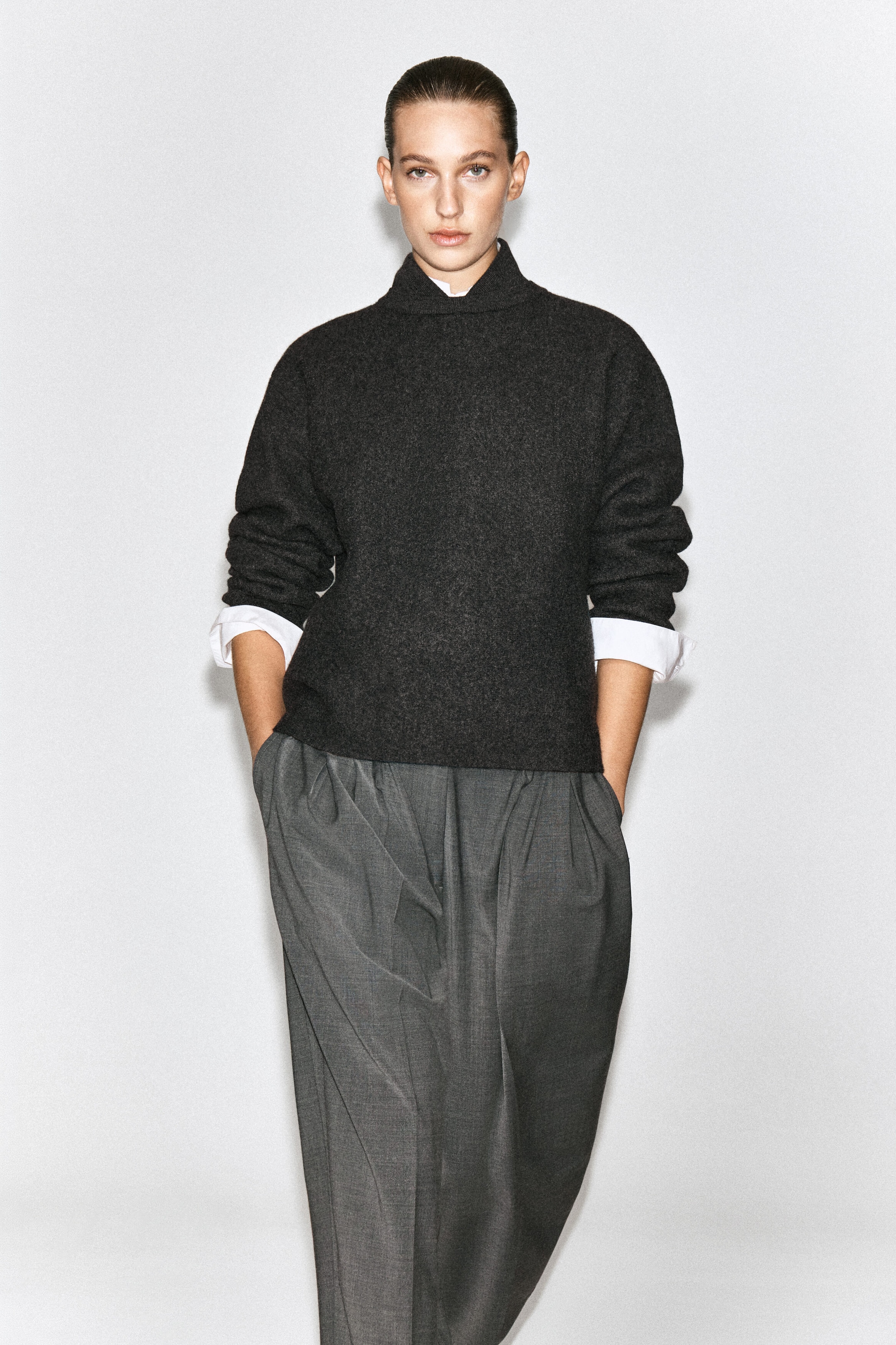 A dark gray boiled wool sweater with a high, layered collar is worn by the model over a white collared shirt. The relaxed-fit sweater features long sleeves with folded cuffs that reveal the white shirt underneath and is paired with gray pleated trousers.