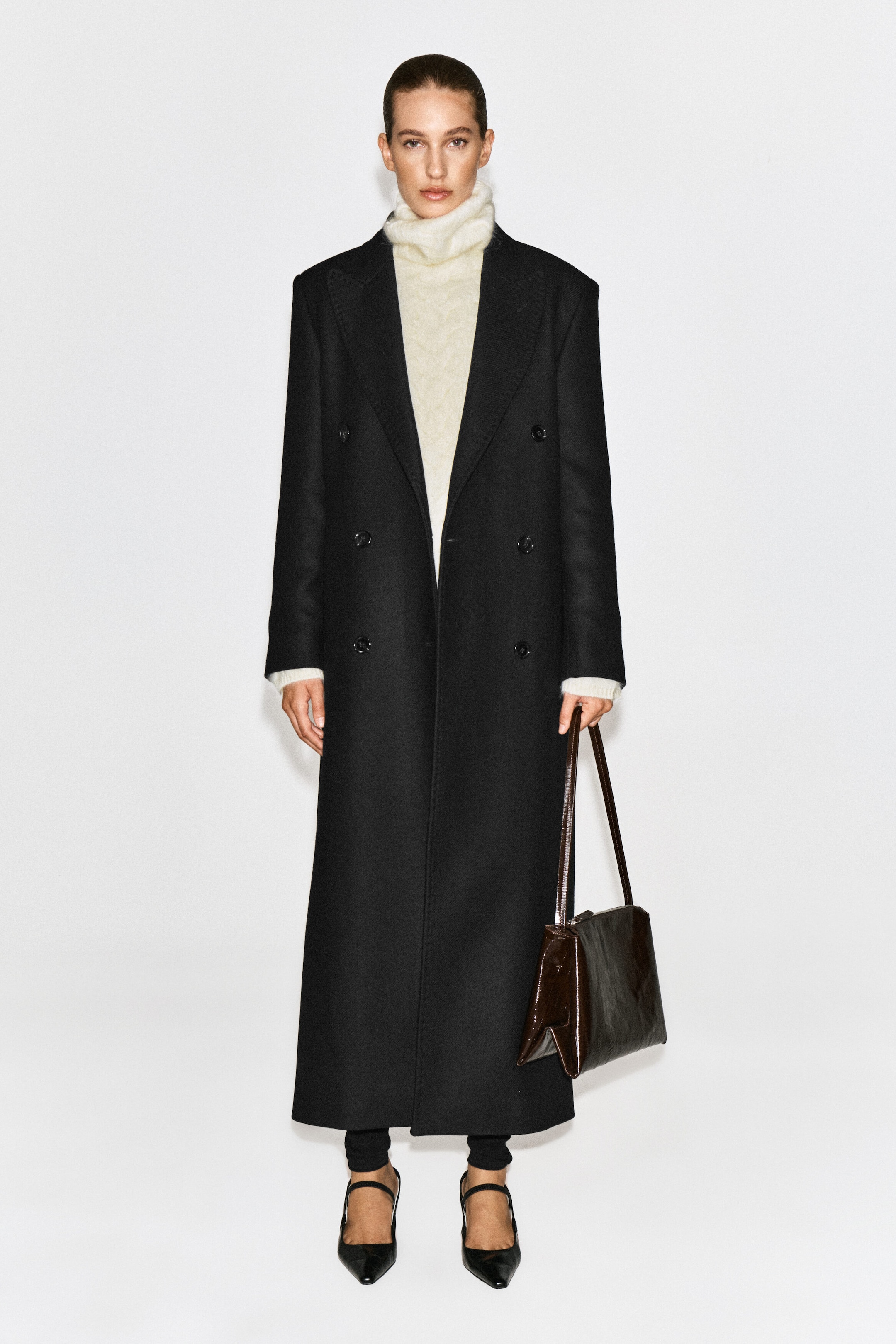 A cream-coloured cable-knit brushed-cashmere funnel-neck jumper is being worn by the model, layered underneath a long, double-breasted black coat. The jumper's high, textured collar is visible above the coat's lapels.