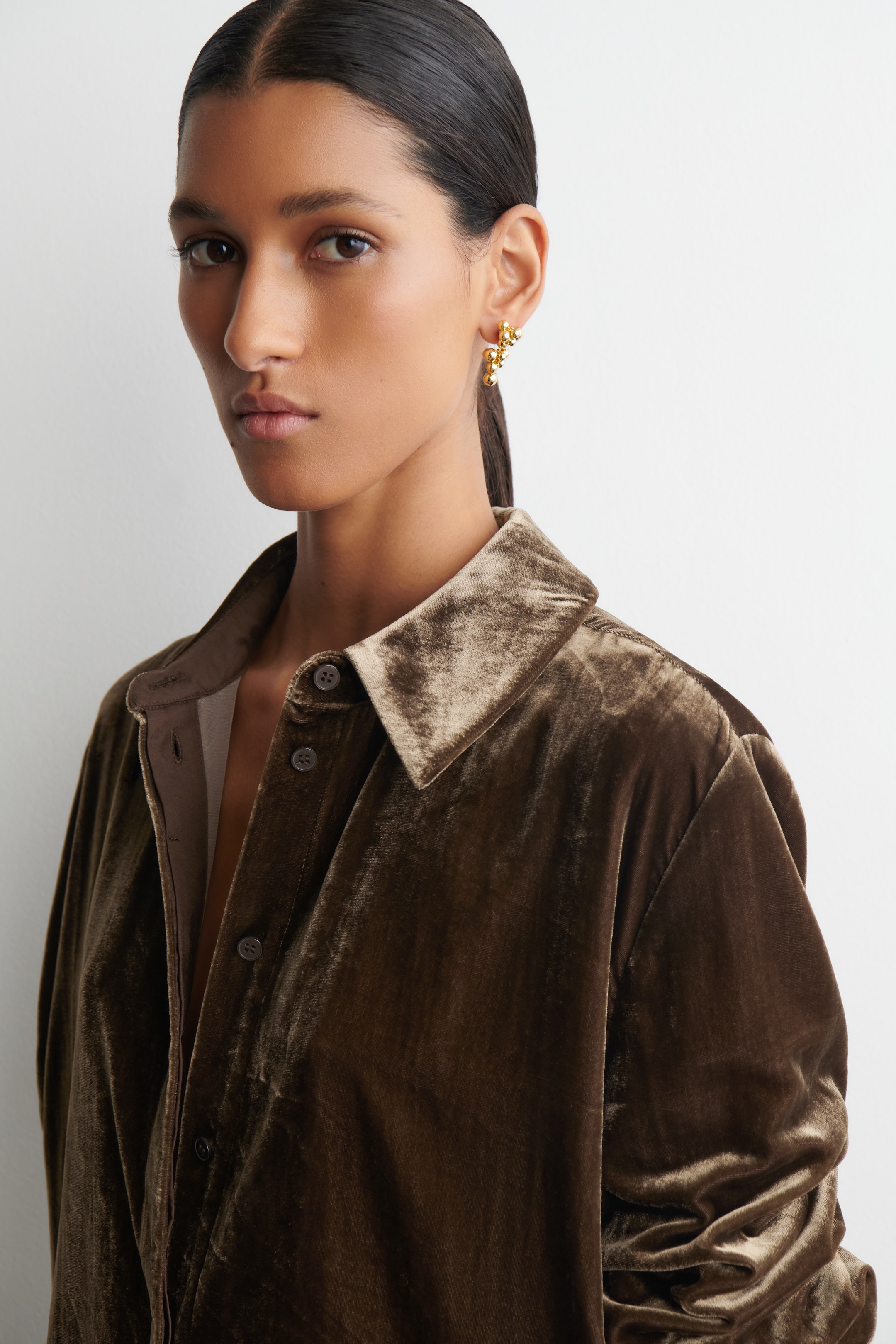 View larger image: An olive-brown velvet shirt is being presented, seen from the front and slightly to the side. It is featuring a classic pointed collar and dark buttons down the front placket, with the long sleeves showing gathered detailing.