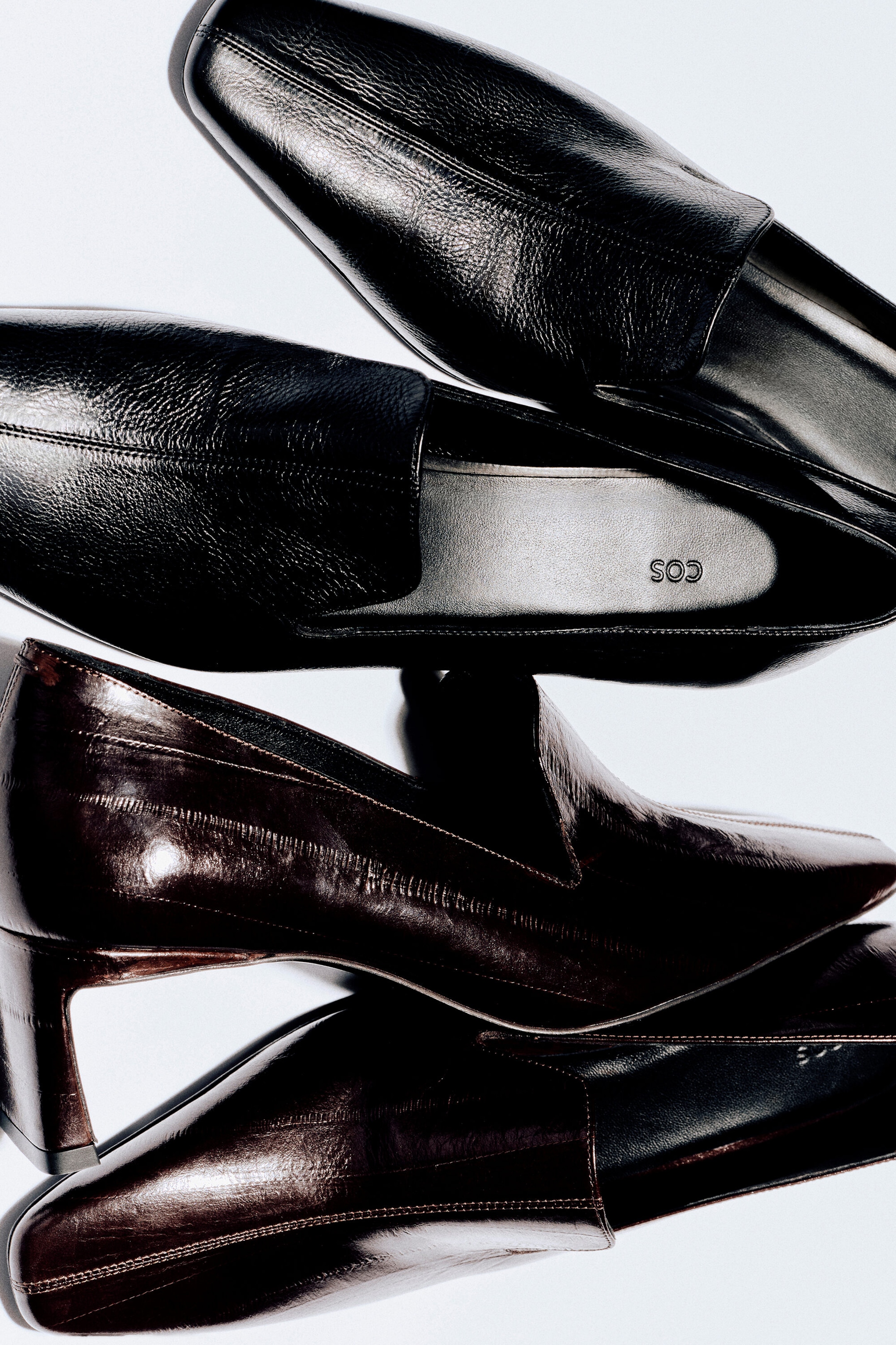 The angular leather heeled loafers are displayed in a stacked arrangement, showcasing one pair in black textured leather and another in glossy dark brown leather. The shoes feature sharp, angular square toes, high-cut vamps, and a low block heel visible in the side profile of the dark brown pair.