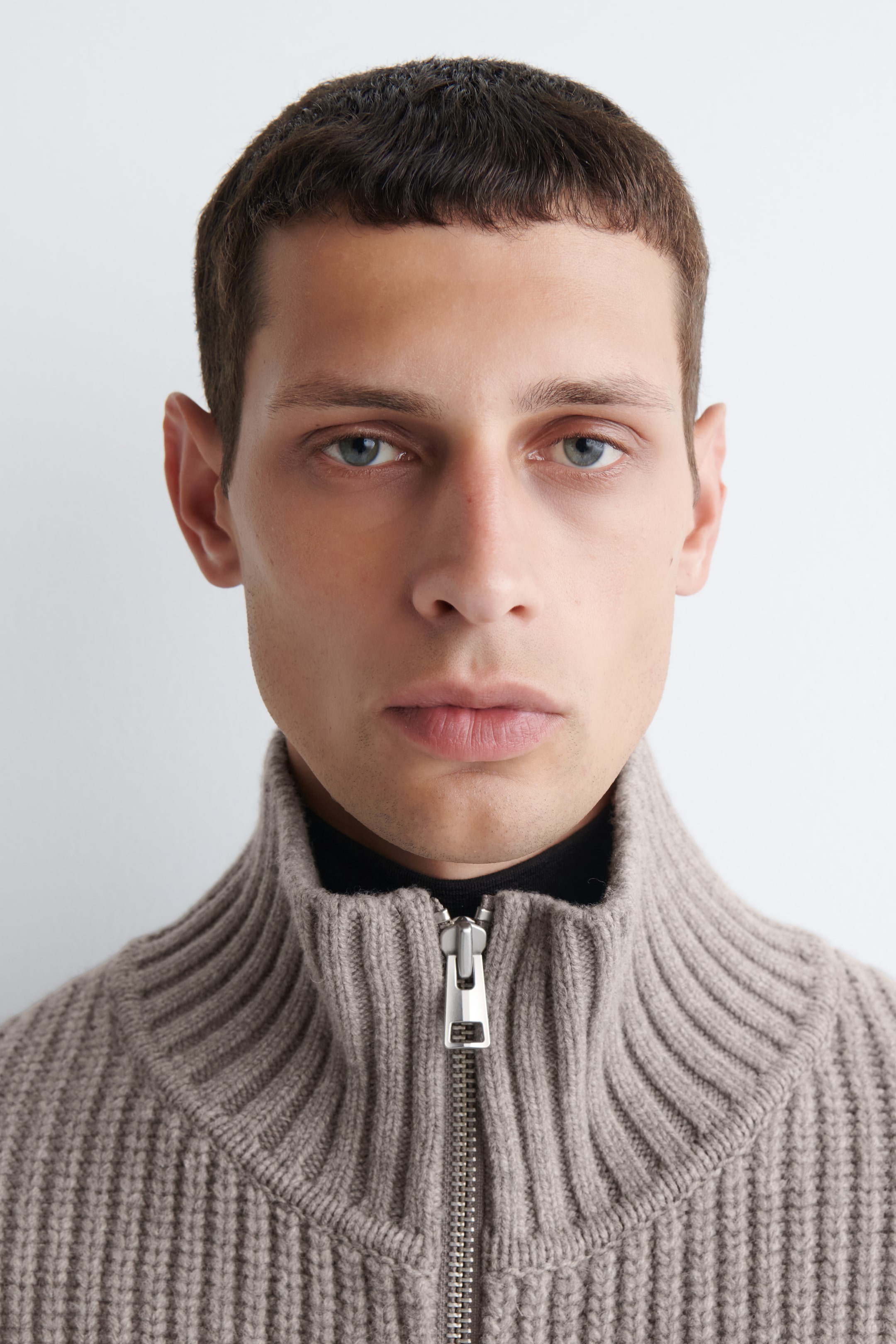 Se større billede: En lysebrun ribstrikket sweater præsenteres fra en frontvendt, close-up vinkel, med en høj stand-up krave og en delvist lynlåset sølvfarvet lynlås. Sweateren fremviser sin fremtrædende lodrette ribstrikkede tekstur og bæres over en sort top, som er synlig ved halsudskæringen.