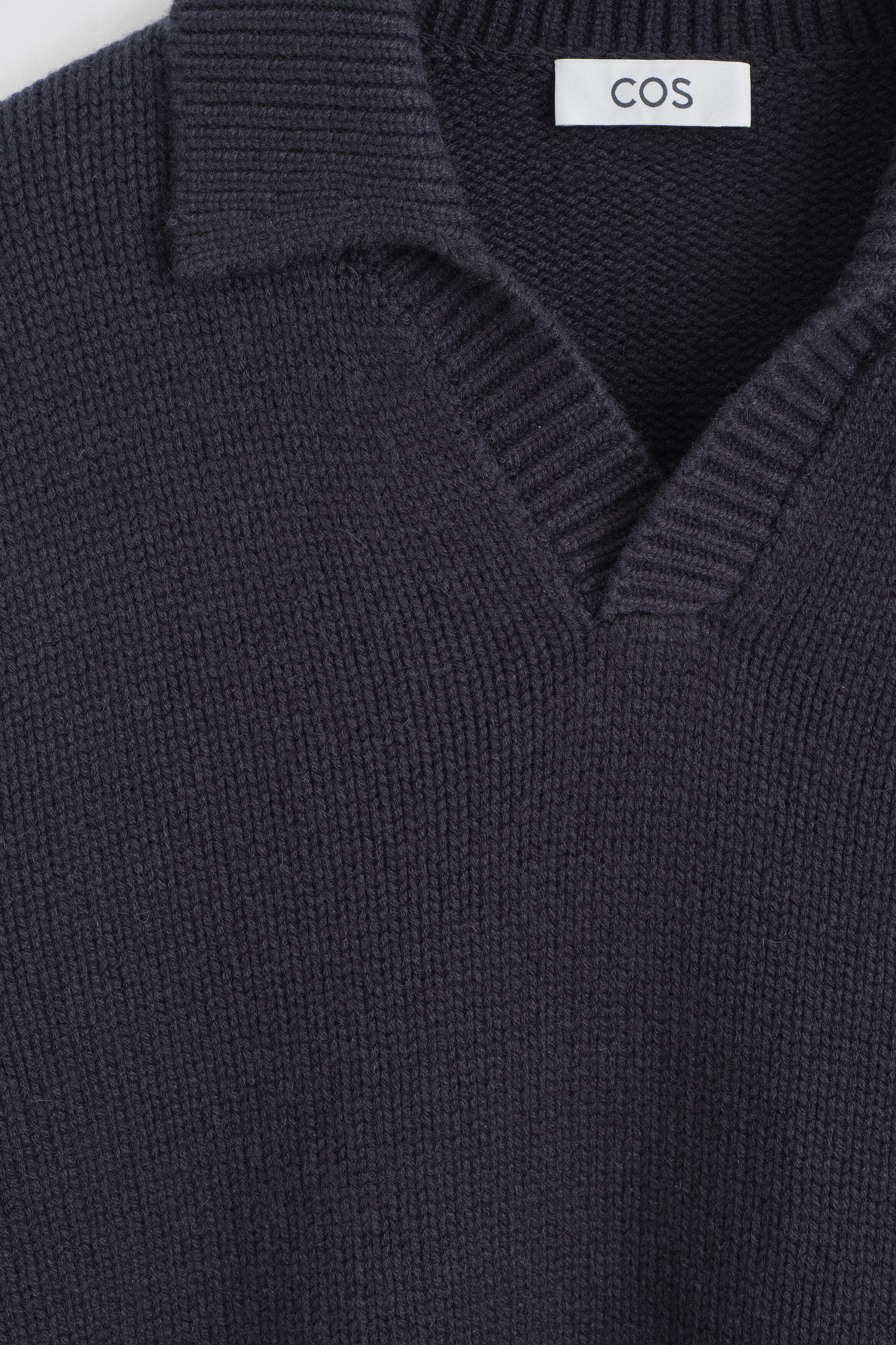 A dark navy blue knit polo sweater features a chunky, textured stitch and a ribbed open collar in a close-up shot. A white brand label with "COS" is visible sewn on the inside back collar. A dark navy blue knit polo sweater features a chunky, textured stitch and a ribbed open collar in a close-up shot. A white brand label with "COS" is visible sewn on the inside back collar.