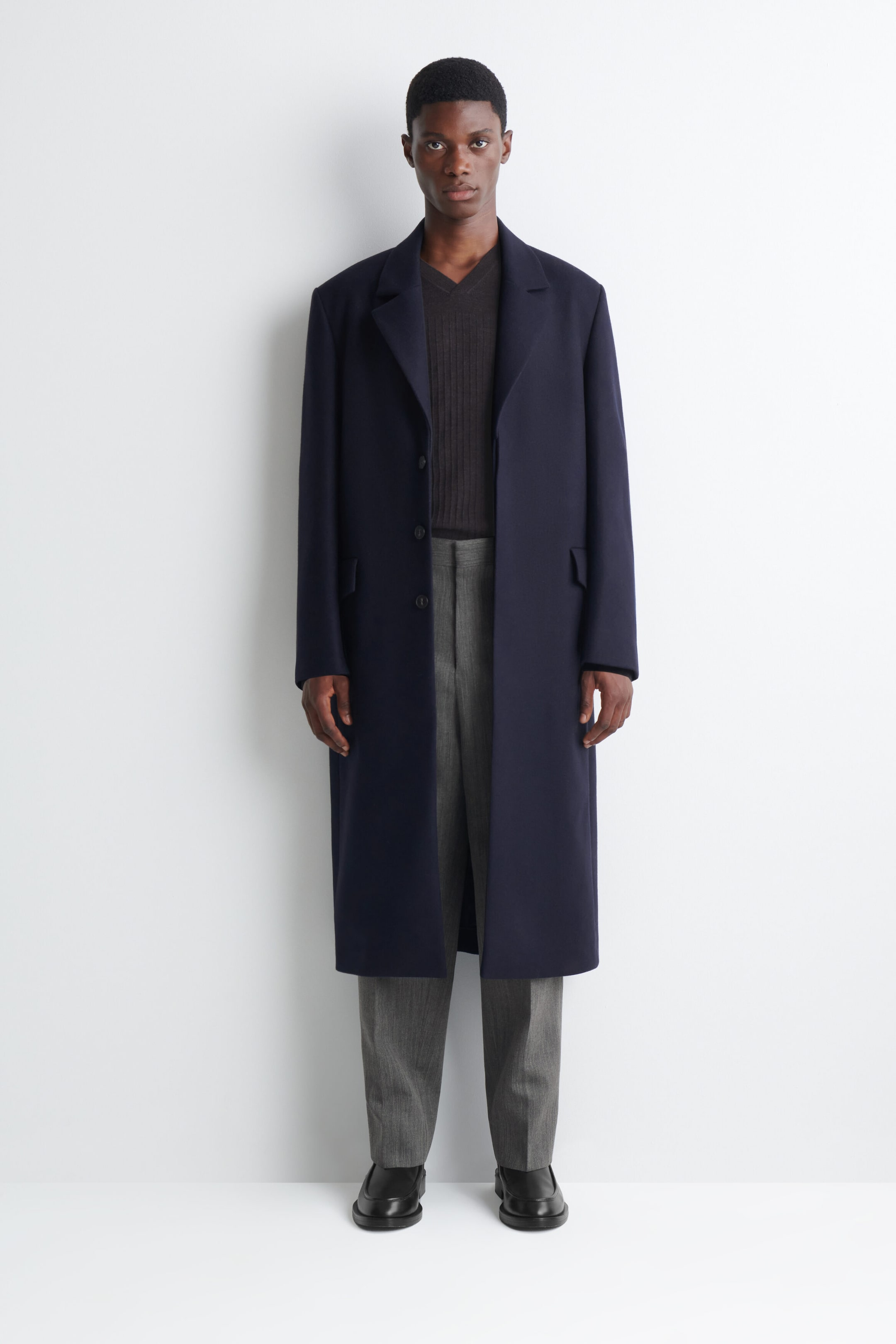 The model is wearing a navy blue single-breasted overcoat. It features notched lapels, three front buttons and two flap pockets, layered over a dark ribbed V-neck jumper and grey trousers. The model is wearing a navy blue single-breasted overcoat. It features notched lapels, three front buttons and two flap pockets, layered over a dark ribbed V-neck jumper and grey trousers.