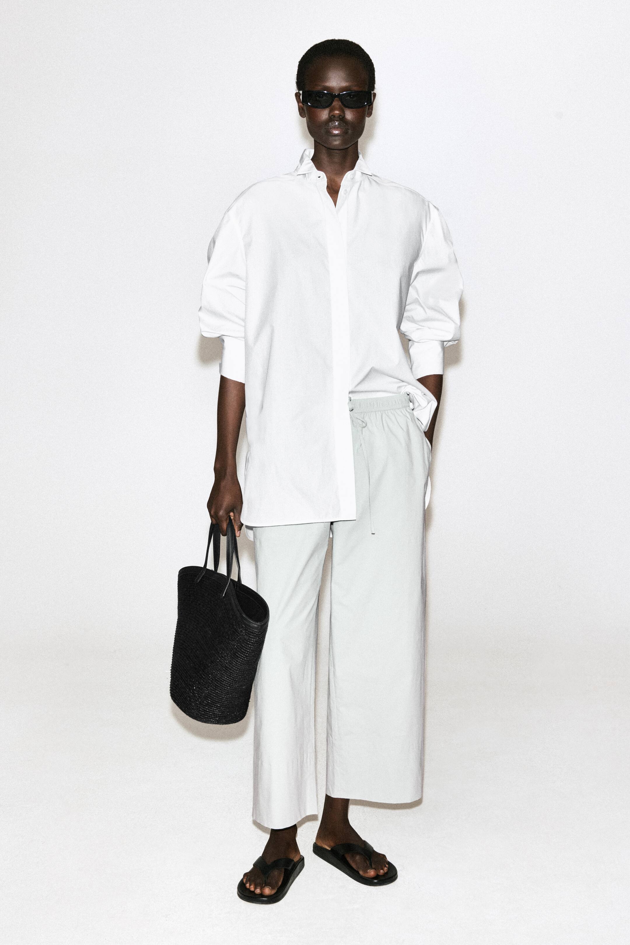 Grotere afbeelding bekijken: Een witte oversized blouse met knoopsluiting, een opstaande kraag en brede manchetten wordt gedragen over lichtgrijze wijde broek met een elastische tailleband met trekkoord. Zwarte teenslippers zijn aan de voeten, en een zwarte geweven shopper met twee handvatten wordt gedragen.