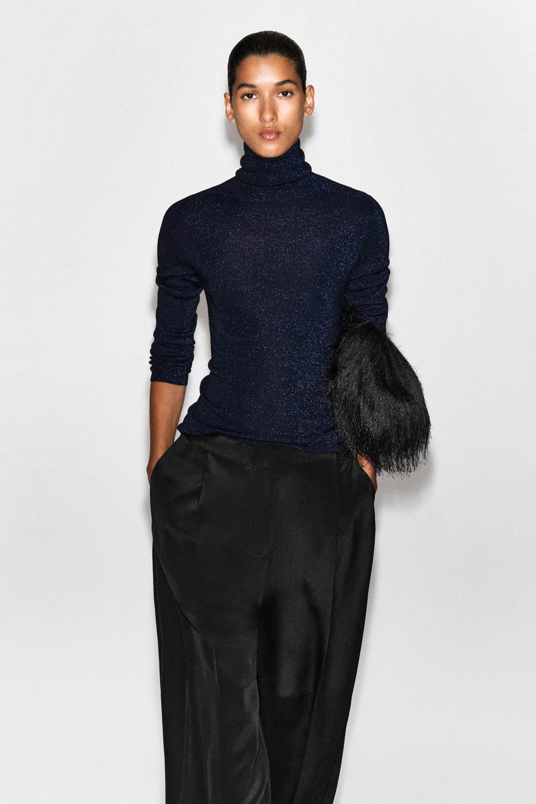 View larger image: A model is showcasing the sparkly seamless wool funnel-neck top from the front, highlighting its slim-fitting silhouette and shimmering dark blue knit fabric. The top features a high, folded funnel neck and long sleeves, styled untucked over black trousers.