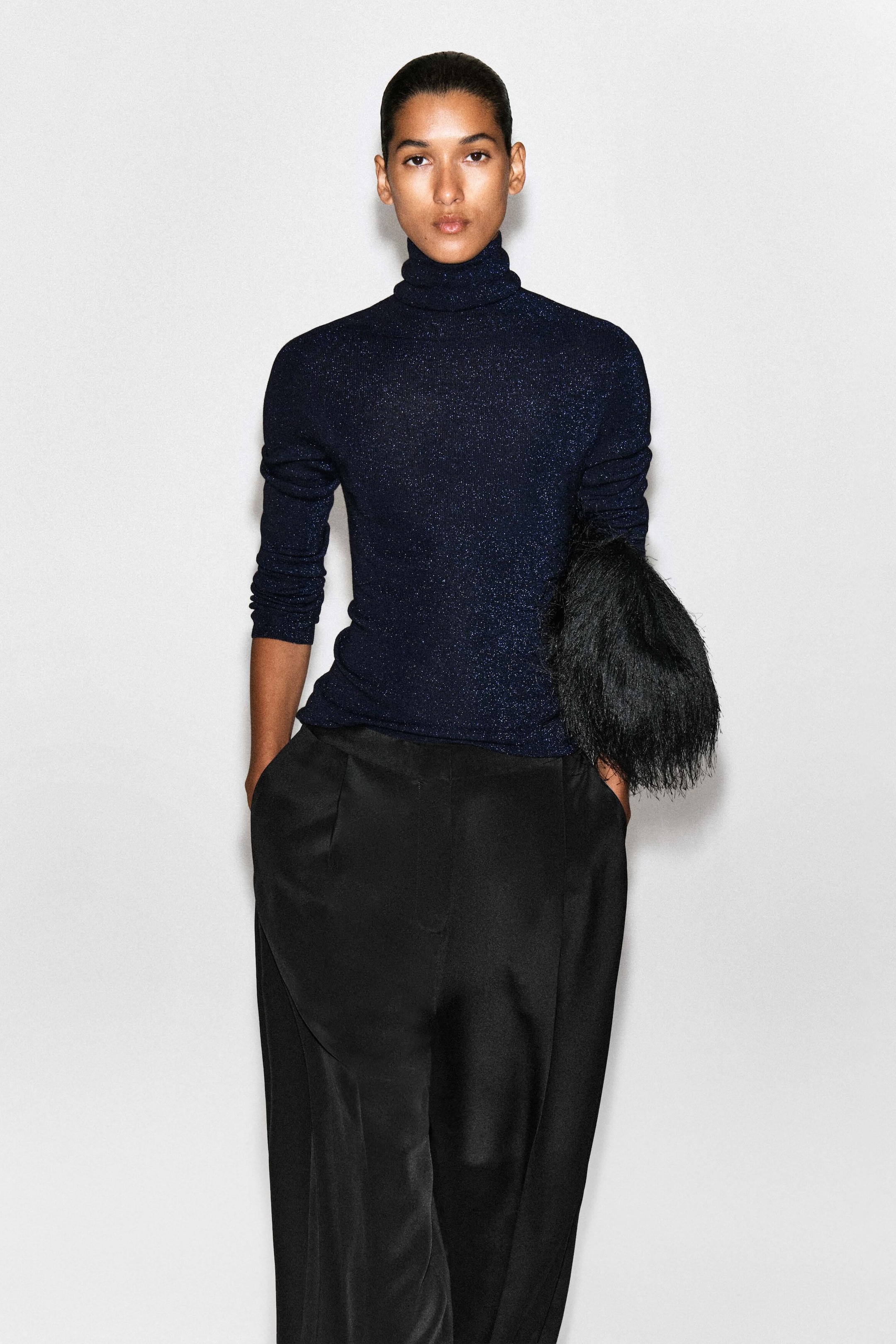A model showcases the sparkly seamless wool funnel-neck top in a front-facing view, highlighting its slim-fitting silhouette and shimmering dark blue knit fabric. The top features a high, folded funnel neck and long sleeves, styled untucked over black trousers.