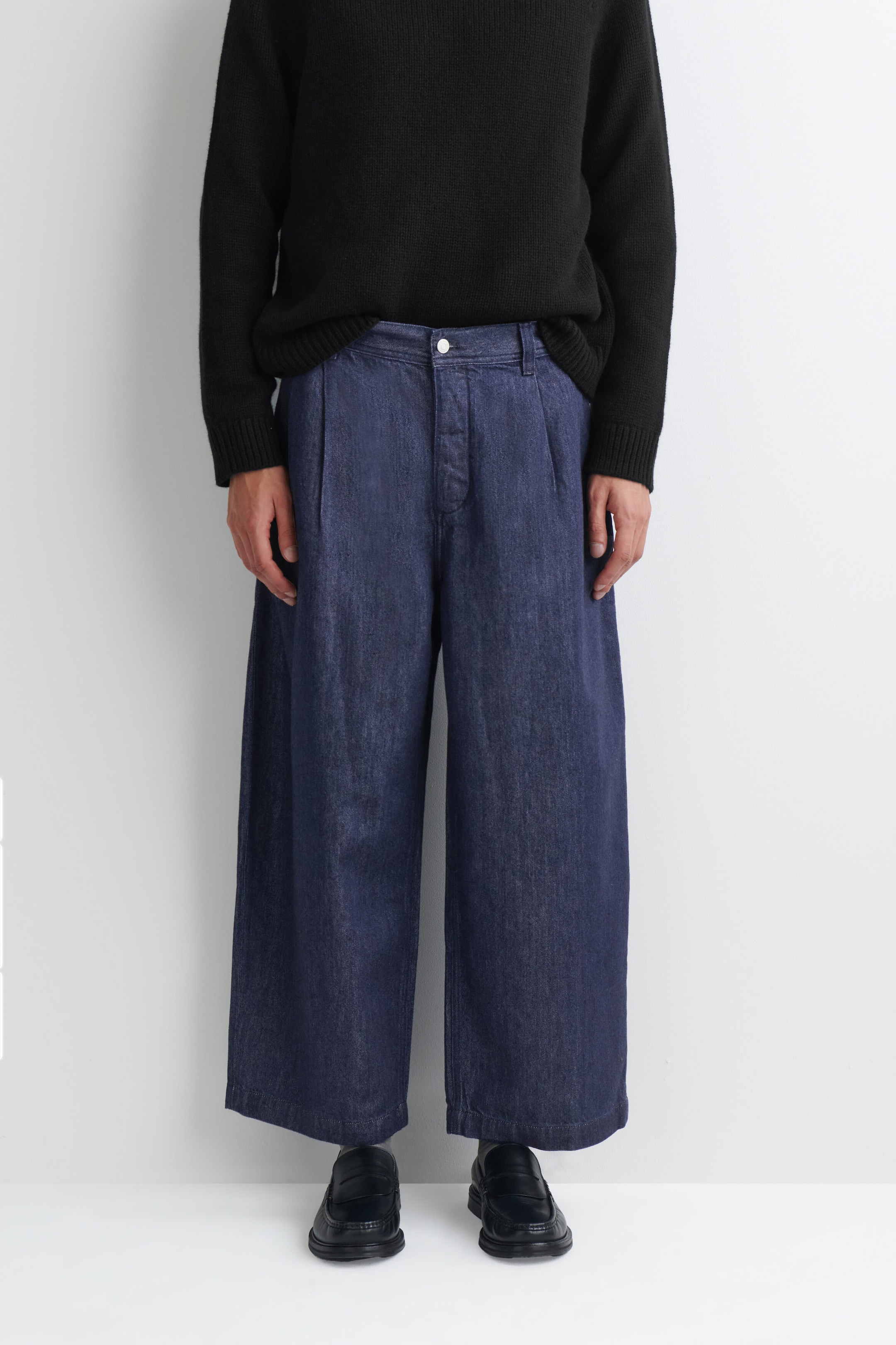 Ce pantalon barrel en denim plissé coupe décontractée, en bleu foncé, est présenté de face, avec une silhouette large et arrondie, des plis sur le devant et une fermeture boutonnée. Il est porté avec un pull en maille noir et des mocassins en cuir noir, mettant en valeur sa coupe décontractée à la longueur cheville. Ce pantalon barrel en denim plissé coupe décontractée, en bleu foncé, est présenté de face, avec une silhouette large et arrondie, des plis sur le devant et une fermeture boutonnée. Il est porté avec un pull en maille noir et des mocassins en cuir noir, mettant en valeur sa coupe décontractée à la longueur cheville.