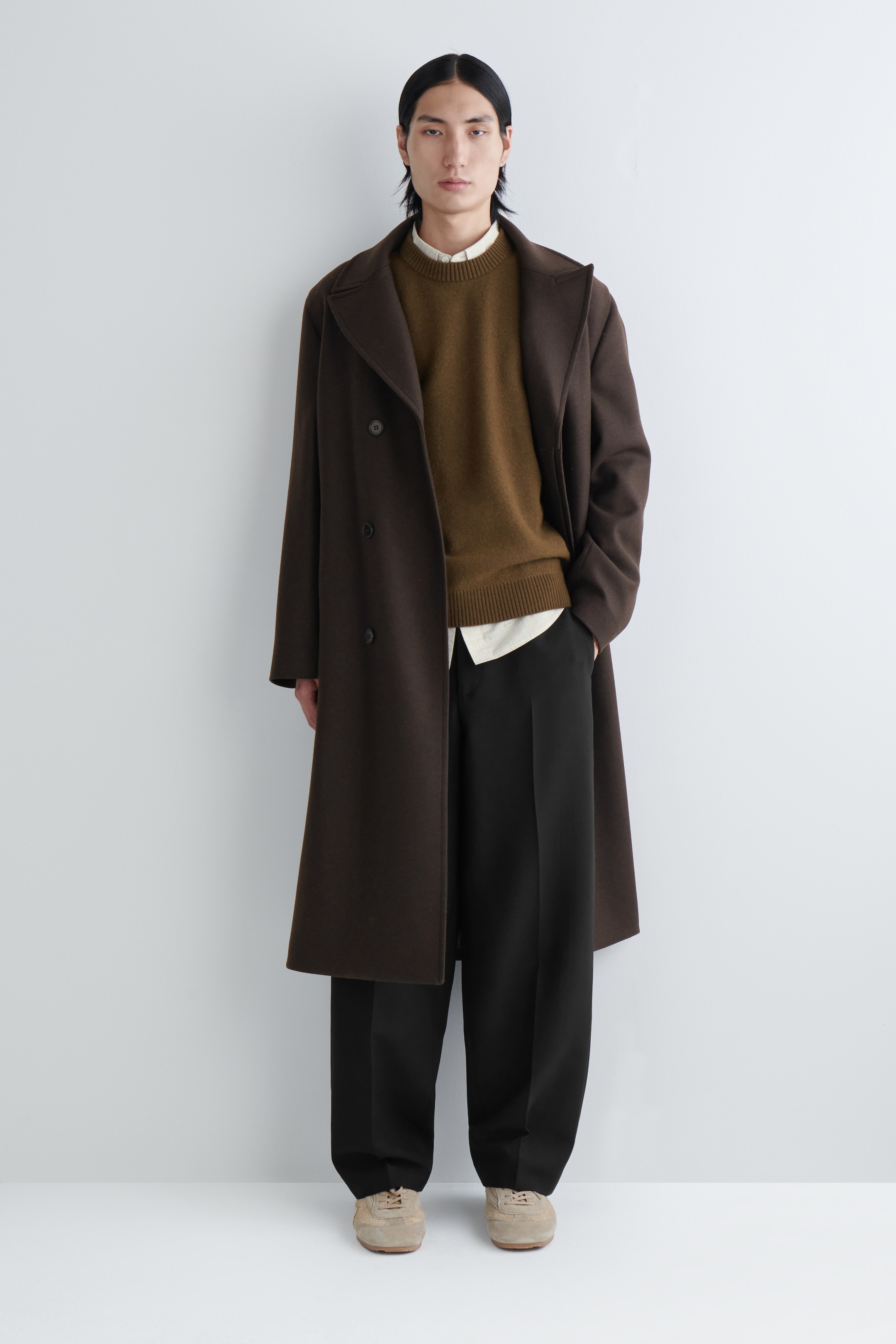 View larger image: A dark brown double-breasted coat with notch lapels is being worn open, revealing a brown crew-neck sweater layered over a light collared shirt. Black wide-leg trousers and light beige sneakers are completing the ensemble.