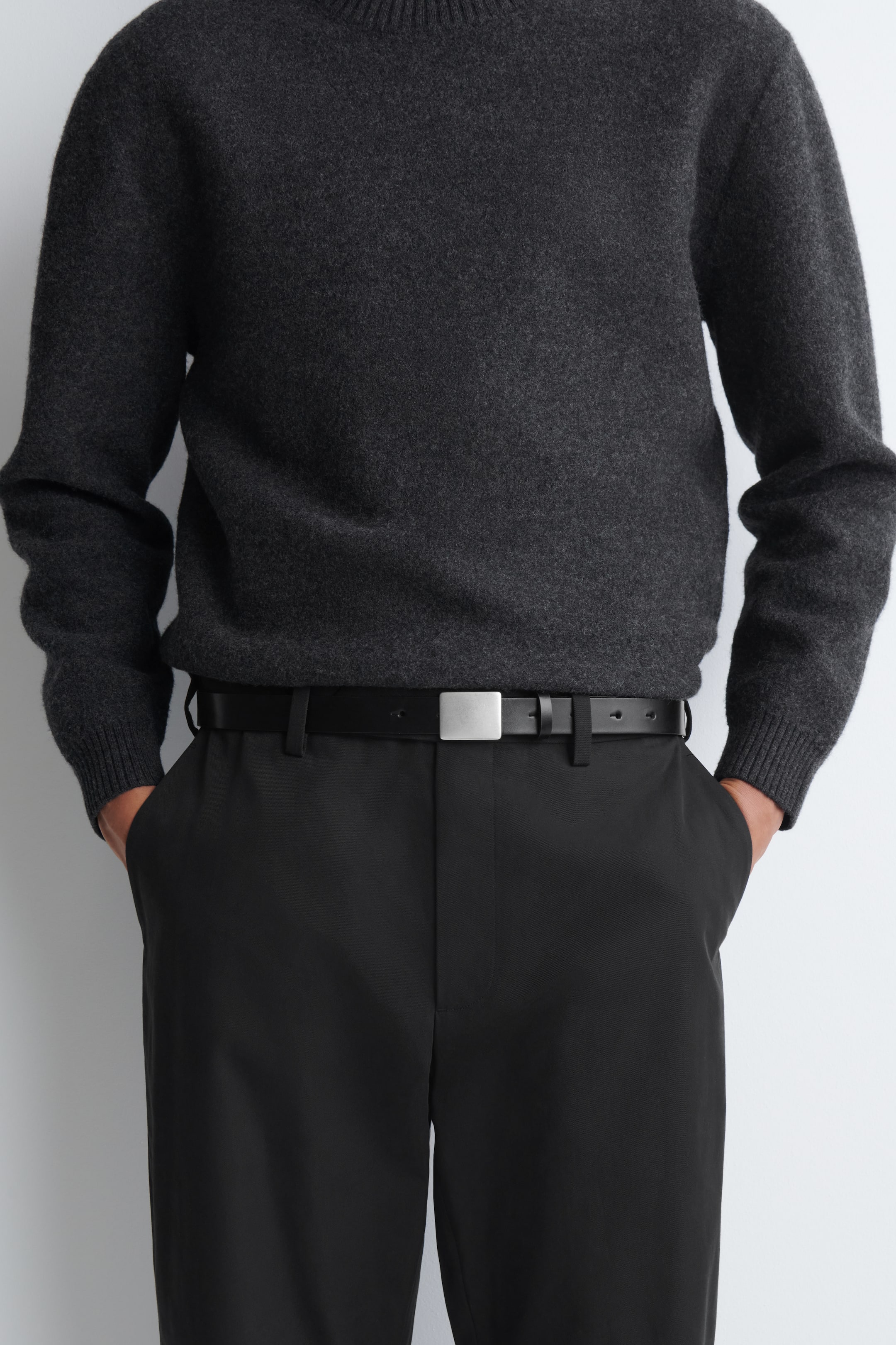 View larger image: A black leather belt featuring a flat, rectangular, matte silver-tone metal buckle is being worn by the model. The belt is threaded through the belt loops of black trousers and styled with a tucked-in dark grey knit sweater.