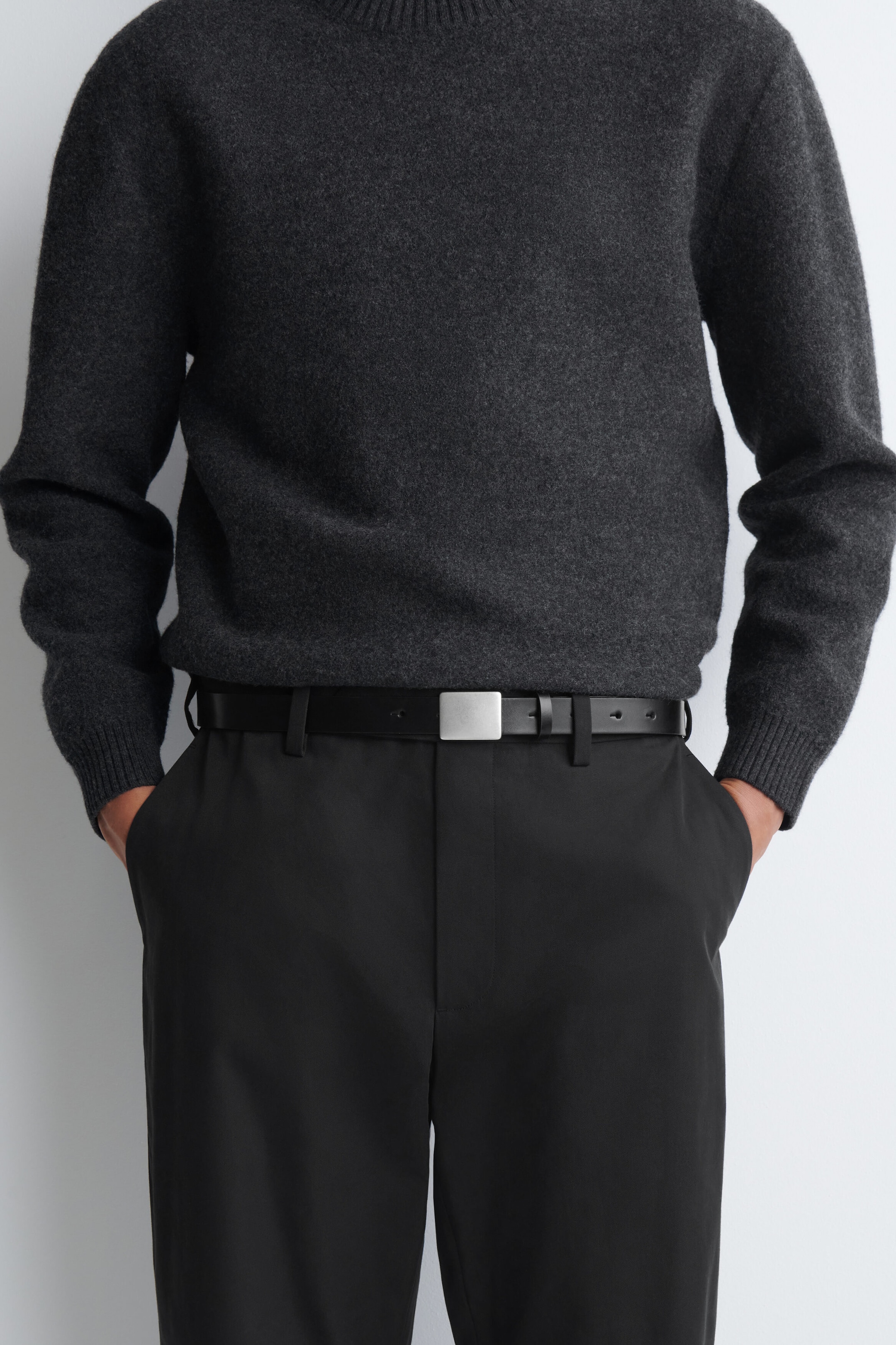 A black leather belt featuring a flat, rectangular, matte silver-tone metal buckle is being worn by the model. The belt is threaded through the belt loops of black trousers and styled with a tucked-in dark grey knit sweater.