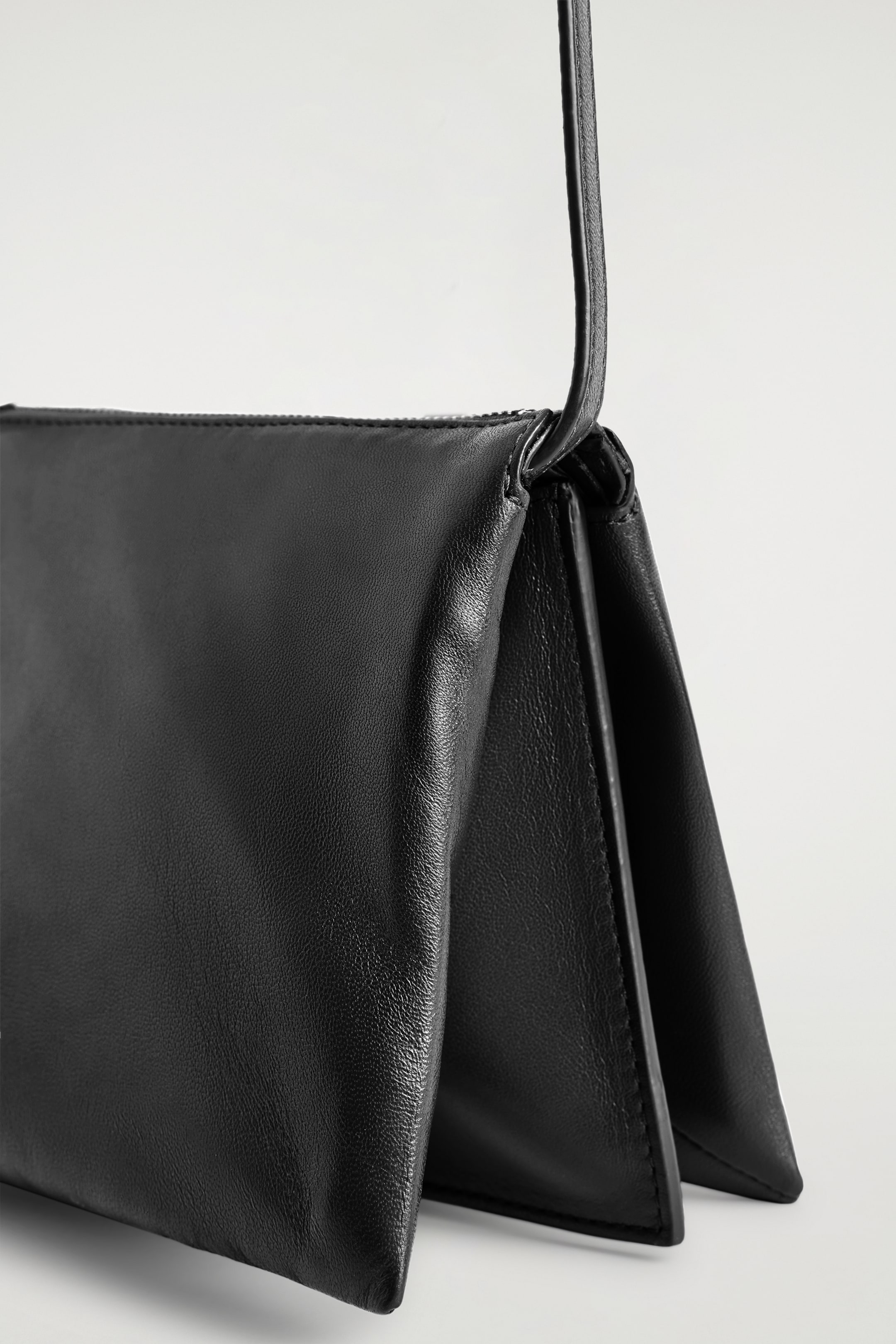 View larger image: TROVE CROSSBODY BAG - LEATHER. This black leather crossbody bag is showcasing a smooth, subtly textured surface and a structured, folded side design, clearly showing its depth as it is standing on its bottom. A thin strap is extending from its top corner, and a zipper closure is visible along the top edge.