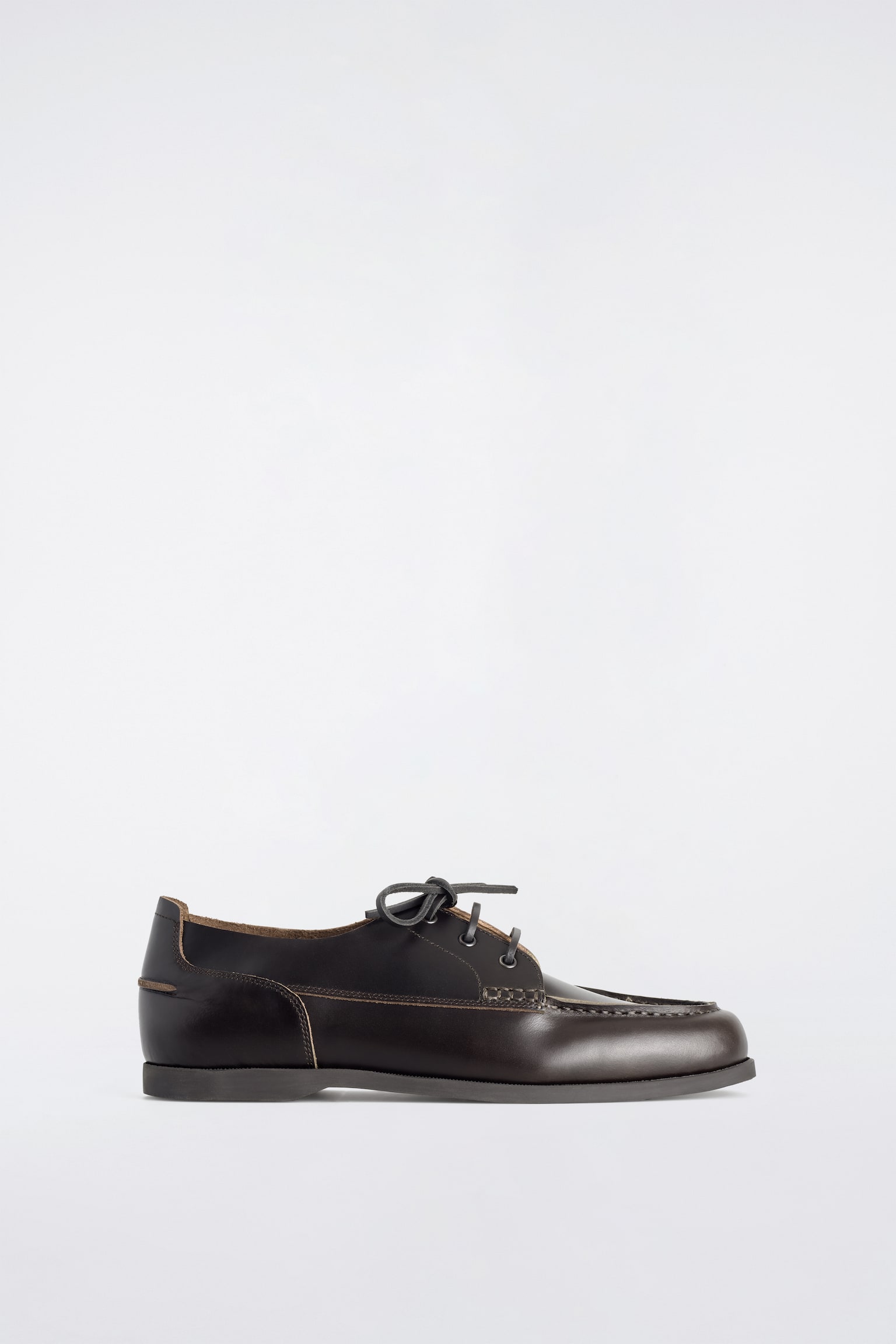 Un zapato de cuero marrón oscuro con cordones está orientado hacia la derecha, con una textura lisa y una construcción moc-toe con costuras marrón claro en el empeine, mientras que los cordones gris oscuro están atados a través de cuatro pares de ojales. - 1
