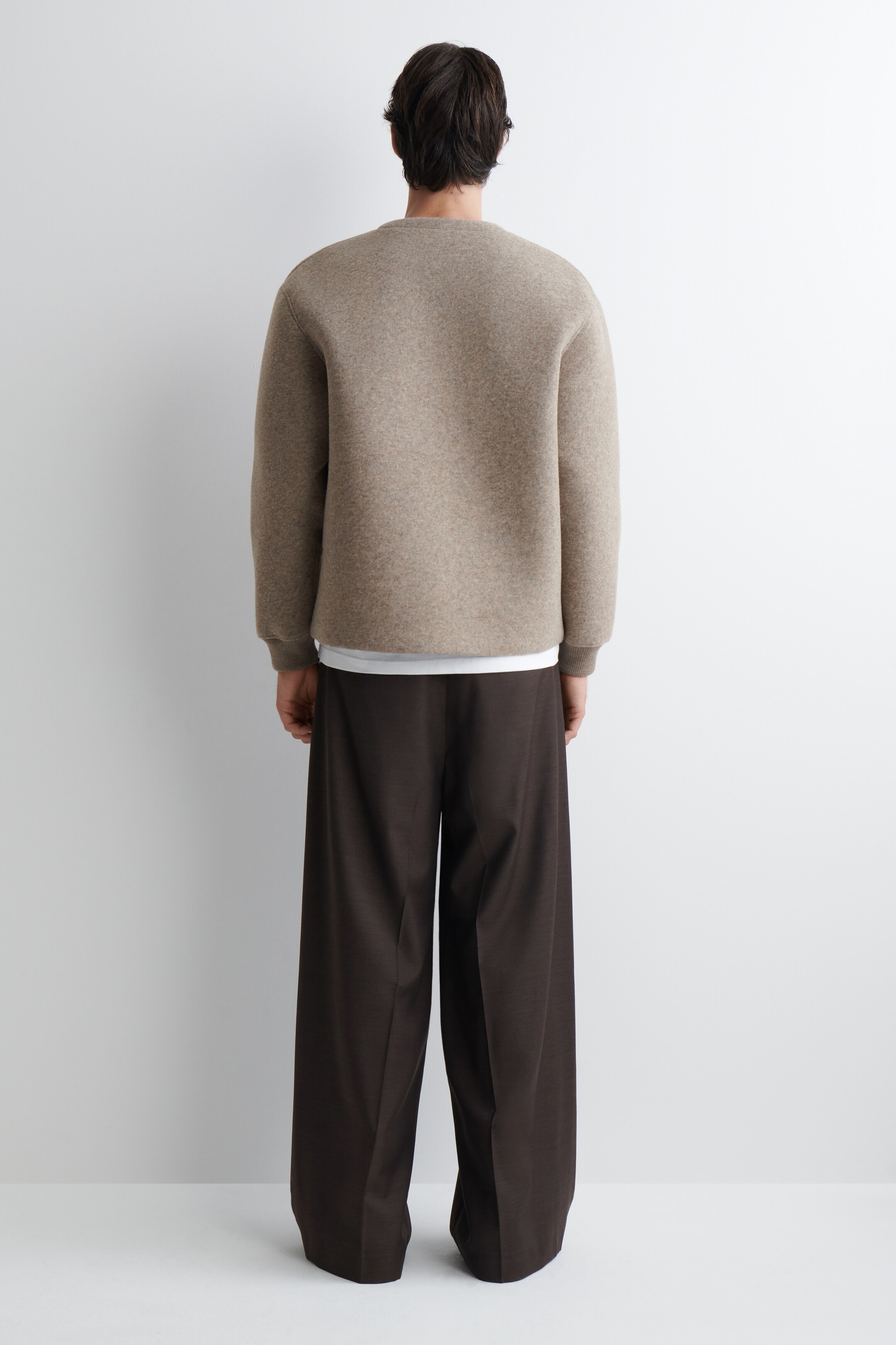 Agrandir l'image: Un pull à col rond brun clair est porté de dos, montrant sa coupe décontractée avec des épaules tombantes et des poignets côtelés, superposé à un t-shirt blanc. Un pantalon large brun foncé complète le look, affichant sa silhouette ample et fluide sous le même angle.