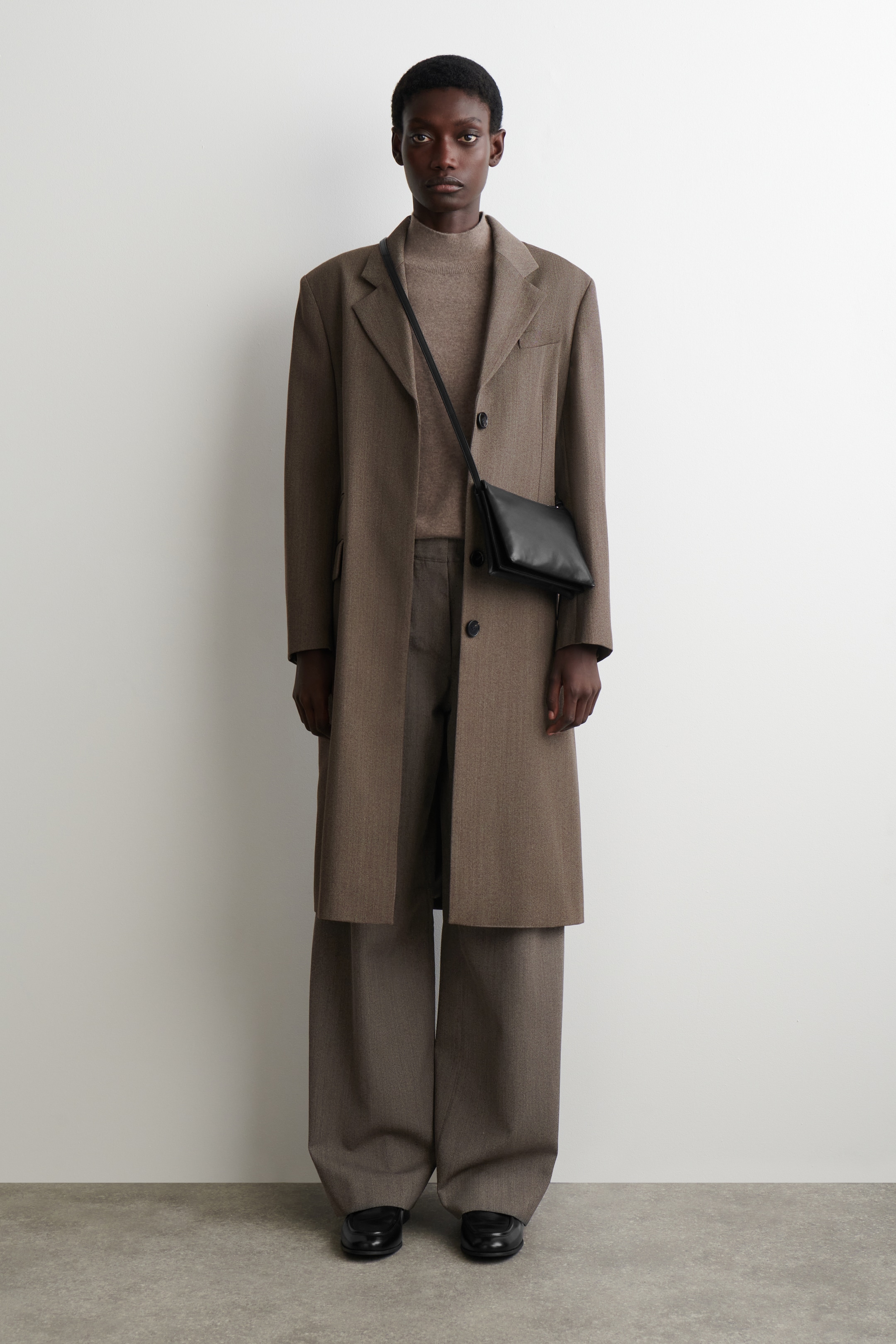 View larger image: A long, single-breasted brown overcoat is being presented from a direct front angle, featuring broad shoulders and notch lapels, and is layered open over a light brown mock-neck sweater and wide-leg trousers. A small black rectangular crossbody bag is being worn across the coat, and black loafers are completing the ensemble.