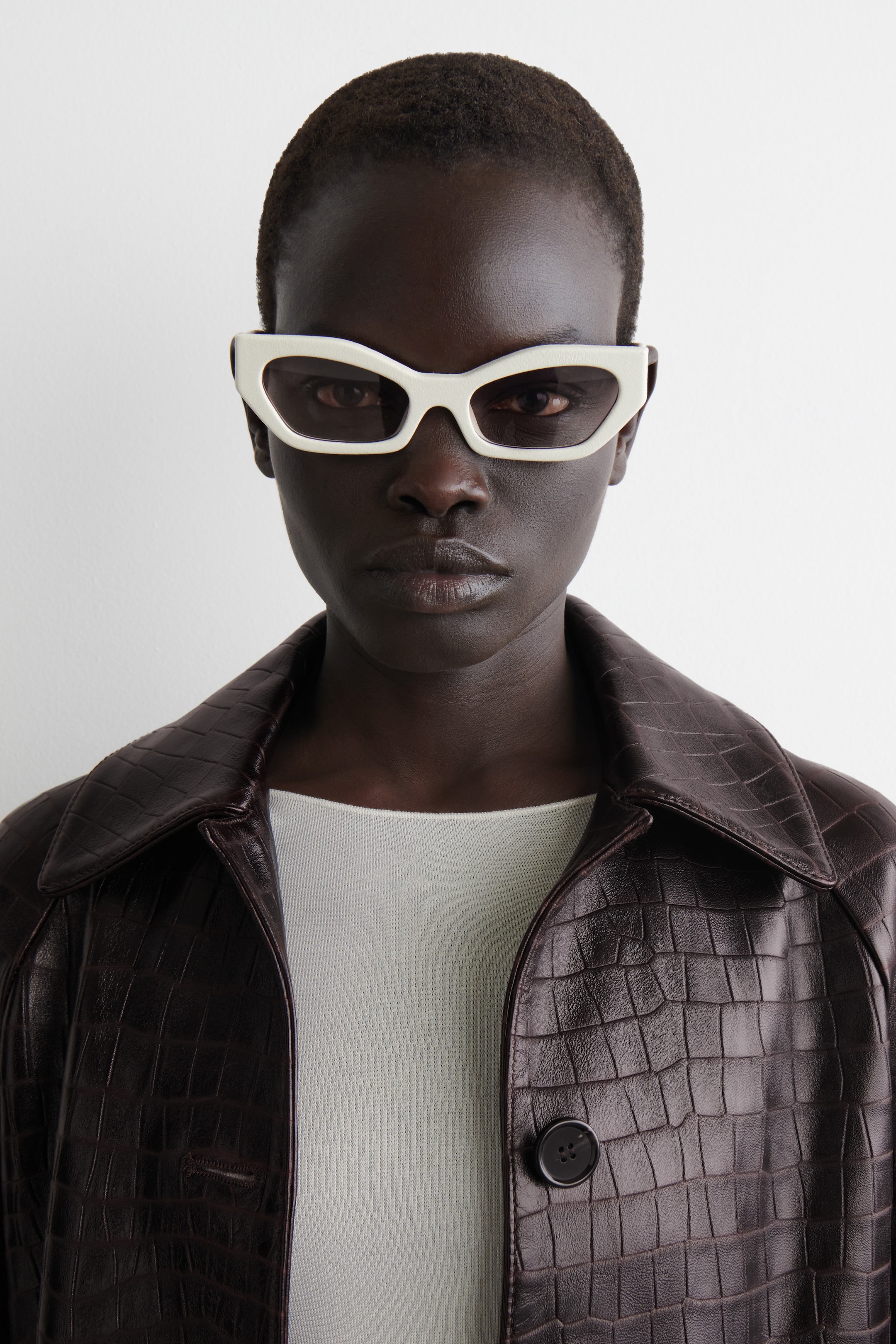 View larger image: Off-white angular cat-eye sunglasses are being worn, featuring dark tinted lenses. A dark brown jacket with a crocodile-embossed texture is being worn open over a light ribbed top, showcasing its pointed collar and a visible front button closure.