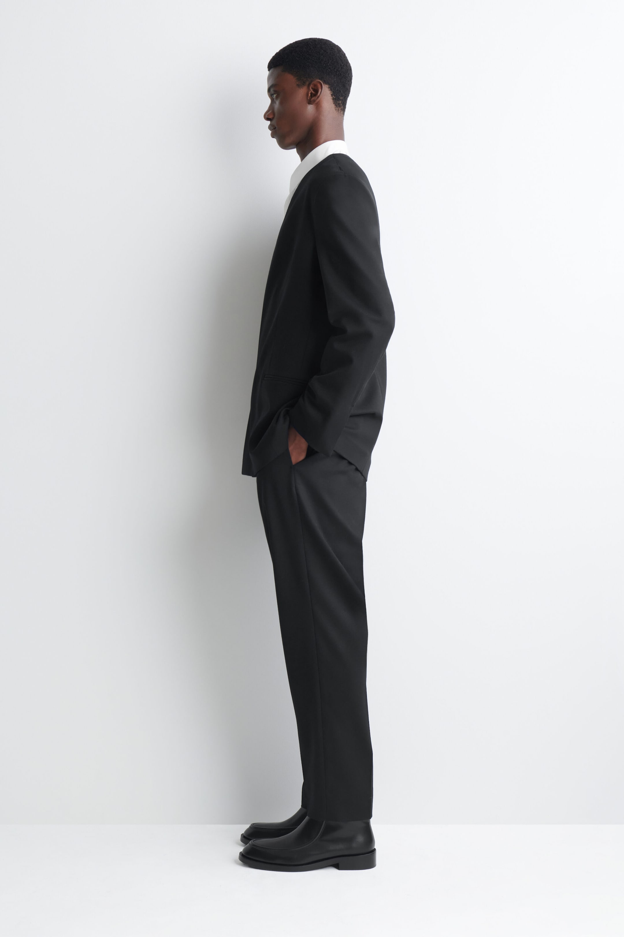 The model, shown in profile, is wearing a black collarless blazer with matching straight-leg trousers. The suit is styled over a white collared shirt, and one hand is resting in the trouser pocket. The model, shown in profile, is wearing a black collarless blazer with matching straight-leg trousers. The suit is styled over a white collared shirt, and one hand is resting in the trouser pocket.
