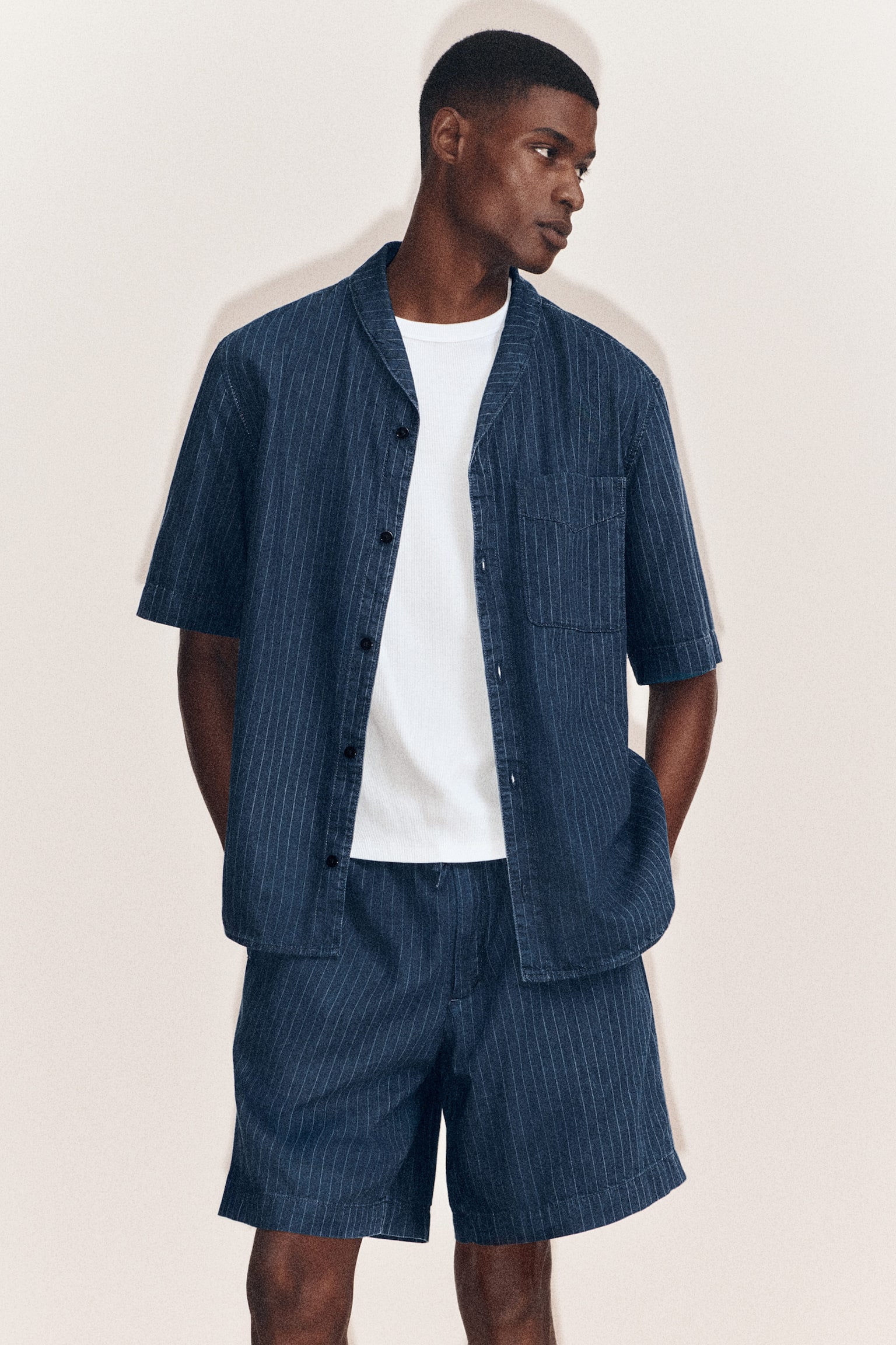 A pinstriped denim shirt and matching pinstriped denim drawstring shorts are worn facing the front, styled with a layered white T-shirt. The short-sleeved shirt features a camp collar, an open button-down front, and a chest patch pocket, while the matching shorts offer a relaxed, coordinated fit. - 1