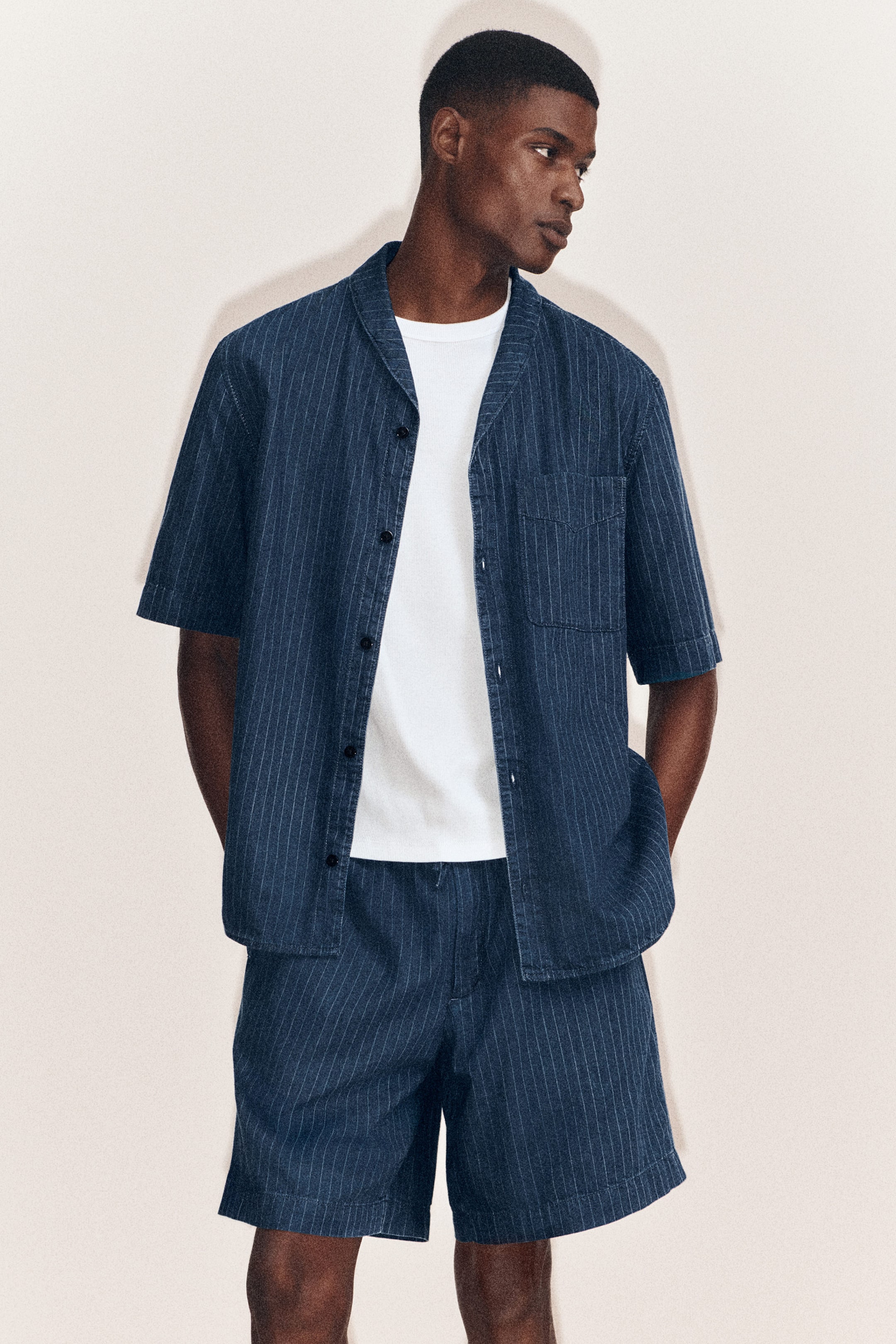 Vezi imaginea mai mare: O cămașă din denim cu dungi fine și pantaloni scurți din denim asortați, cu dungi fine și șnur în talie, sunt purtate frontal, stilizate cu un tricou alb în straturi. Cămașa cu mânecă scurtă evidențiază un guler camp, un față cu nasturi lăsată deschisă și un buzunar aplicat la piept, în timp ce pantalonii scurți asortați oferă o croială relaxată, coordonată.