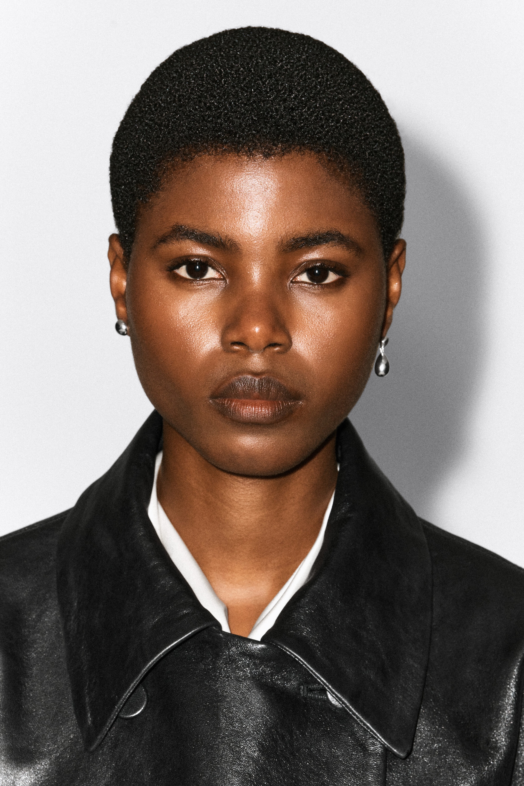 The model is wearing mismatched silver earrings, with a small ball stud on one ear and a polished teardrop drop earring on the other. The jewelry is styled with a black leather jacket over a white shirt.