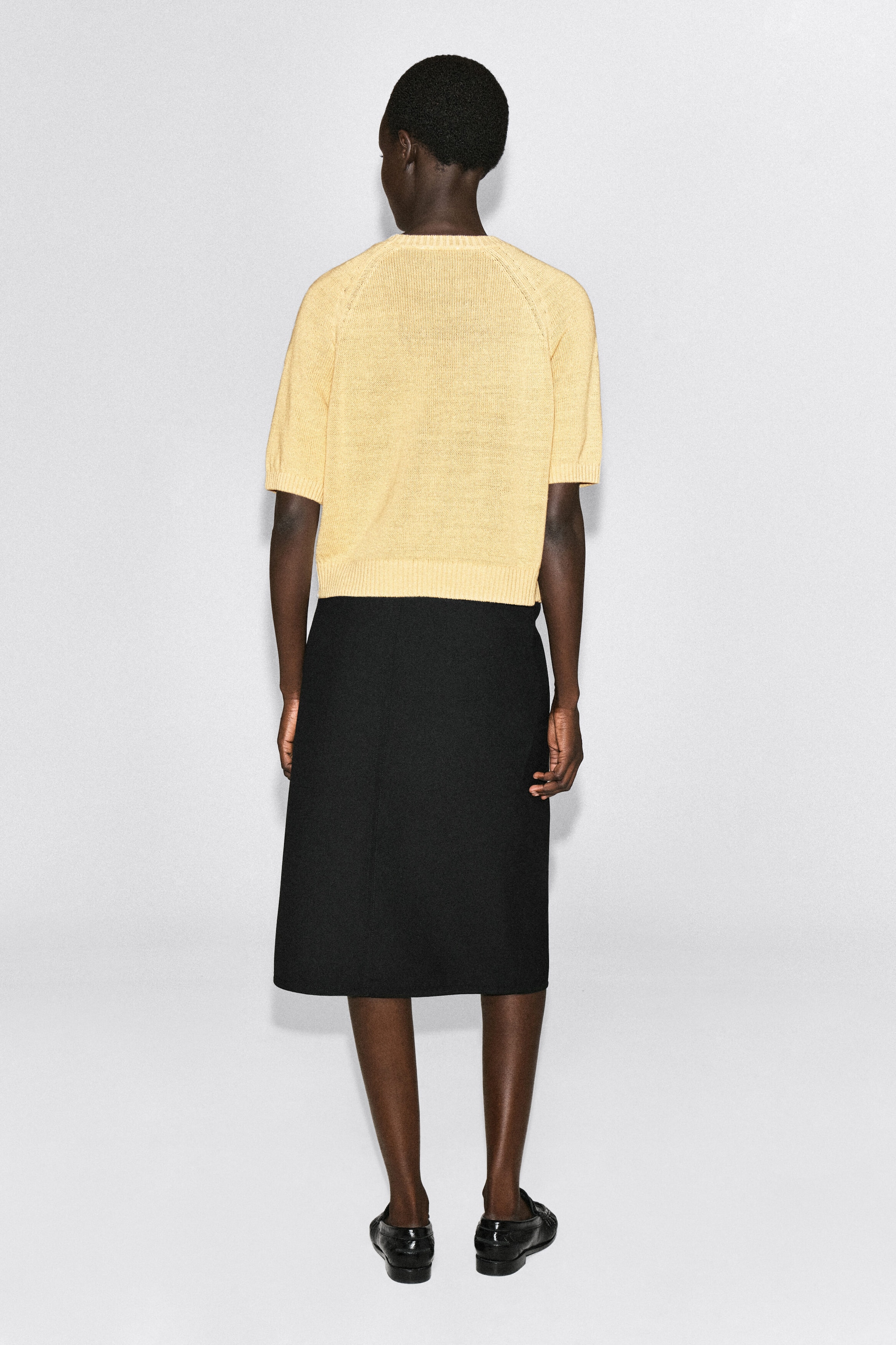 The Shrunken Knitted Linen T-shirt is shown from the back, featuring a pale yellow, lightweight knit with short raglan sleeves, ribbed cuffs, and a ribbed hem. The garment is shown in a cropped fit that sits just above a black midi skirt. The Shrunken Knitted Linen T-shirt is shown from the back, featuring a pale yellow, lightweight knit with short raglan sleeves, ribbed cuffs, and a ribbed hem. The garment is shown in a cropped fit that sits just above a black midi skirt.