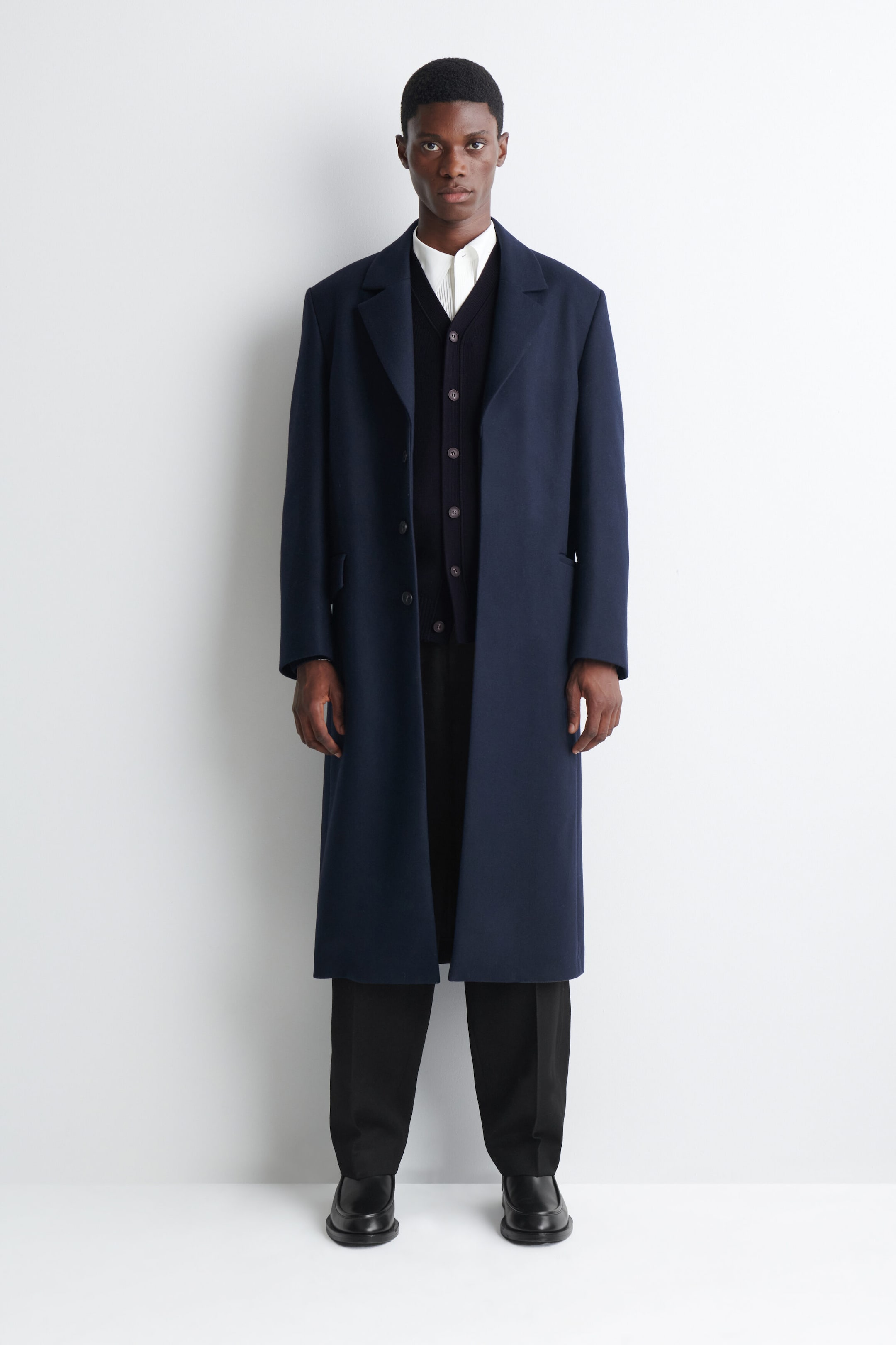 A long navy blue overcoat featuring notch lapels and side pockets is being worn by the model. The open-front coat is layered over a dark buttoned cardigan, a white collared shirt, and black trousers. A long navy blue overcoat featuring notch lapels and side pockets is being worn by the model. The open-front coat is layered over a dark buttoned cardigan, a white collared shirt, and black trousers.