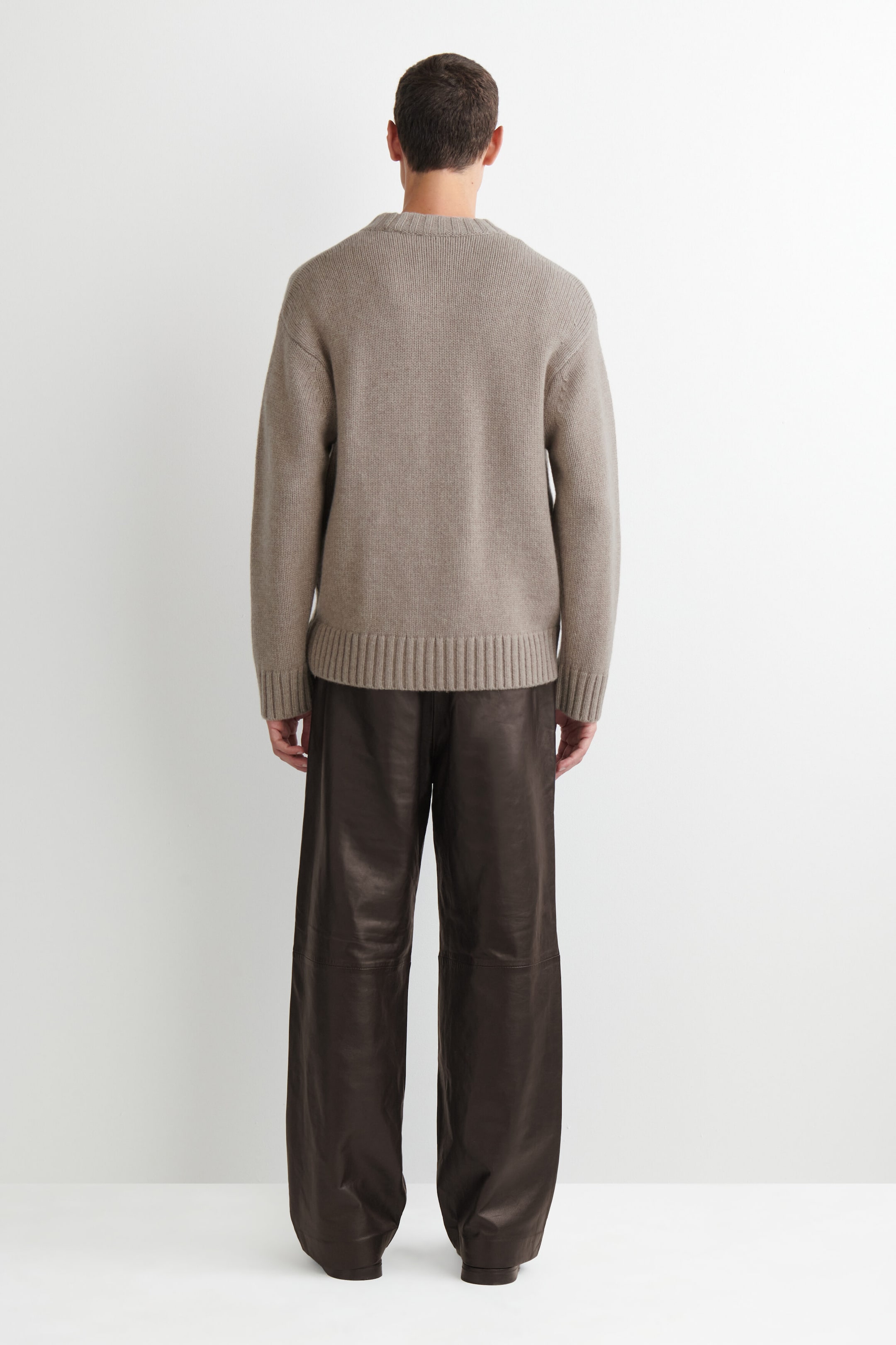 The leather relaxed-fit straight-leg pants are shown from the back in a dark brown colour, draping in a loose, straight silhouette. The pants feature a horizontal seam detail across the backs of the knees and extend down to the floor, with the waistband covered by a beige knit sweater. The leather relaxed-fit straight-leg pants are shown from the back in a dark brown colour, draping in a loose, straight silhouette. The pants feature a horizontal seam detail across the backs of the knees and extend down to the floor, with the waistband covered by a beige knit sweater.