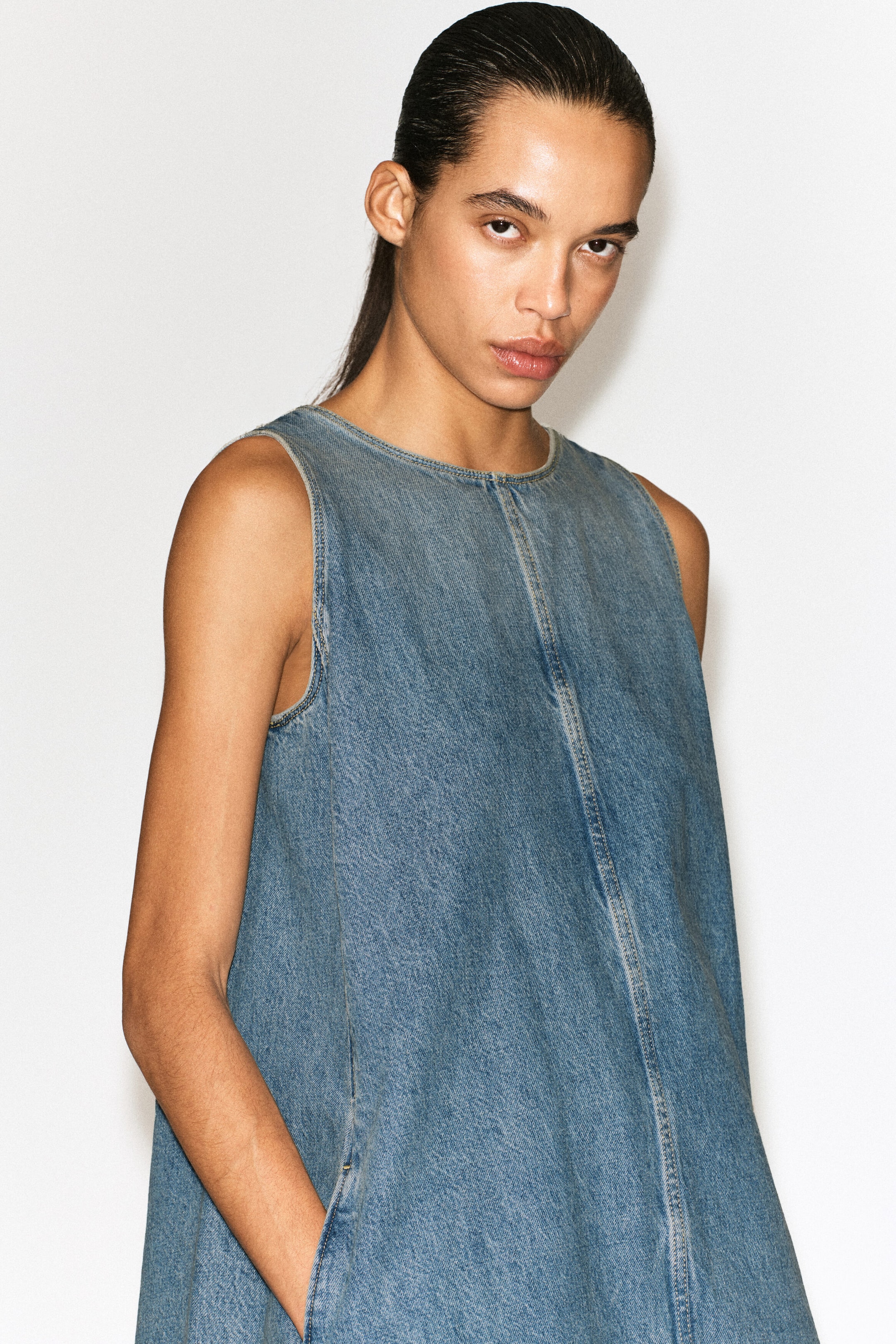Un minidress in denim senza maniche con finitura blu slavata è mostrato frontalmente, caratterizzato da un girocollo e da una vistosa cucitura impunturata centrale sul davanti che definisce la sua silhouette svasata a trapezio rilassata.