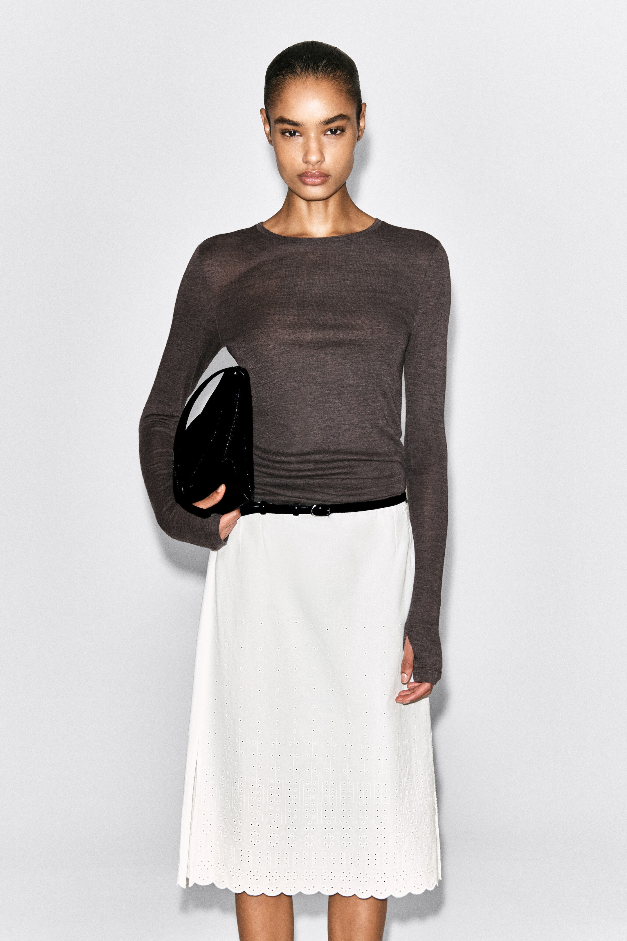 The slim merino wool top is facing front, featuring a dark grey-brown mélange colour, a classic round neckline, and a lightweight, semi-sheer texture. It fits closely to the body with extra-long sleeves extending over the hands, and is tucked into a belted skirt at the waist. The slim merino wool top is facing front, featuring a dark grey-brown mélange colour, a classic round neckline, and a lightweight, semi-sheer texture. It fits closely to the body with extra-long sleeves extending over the hands, and is tucked into a belted skirt at the waist.