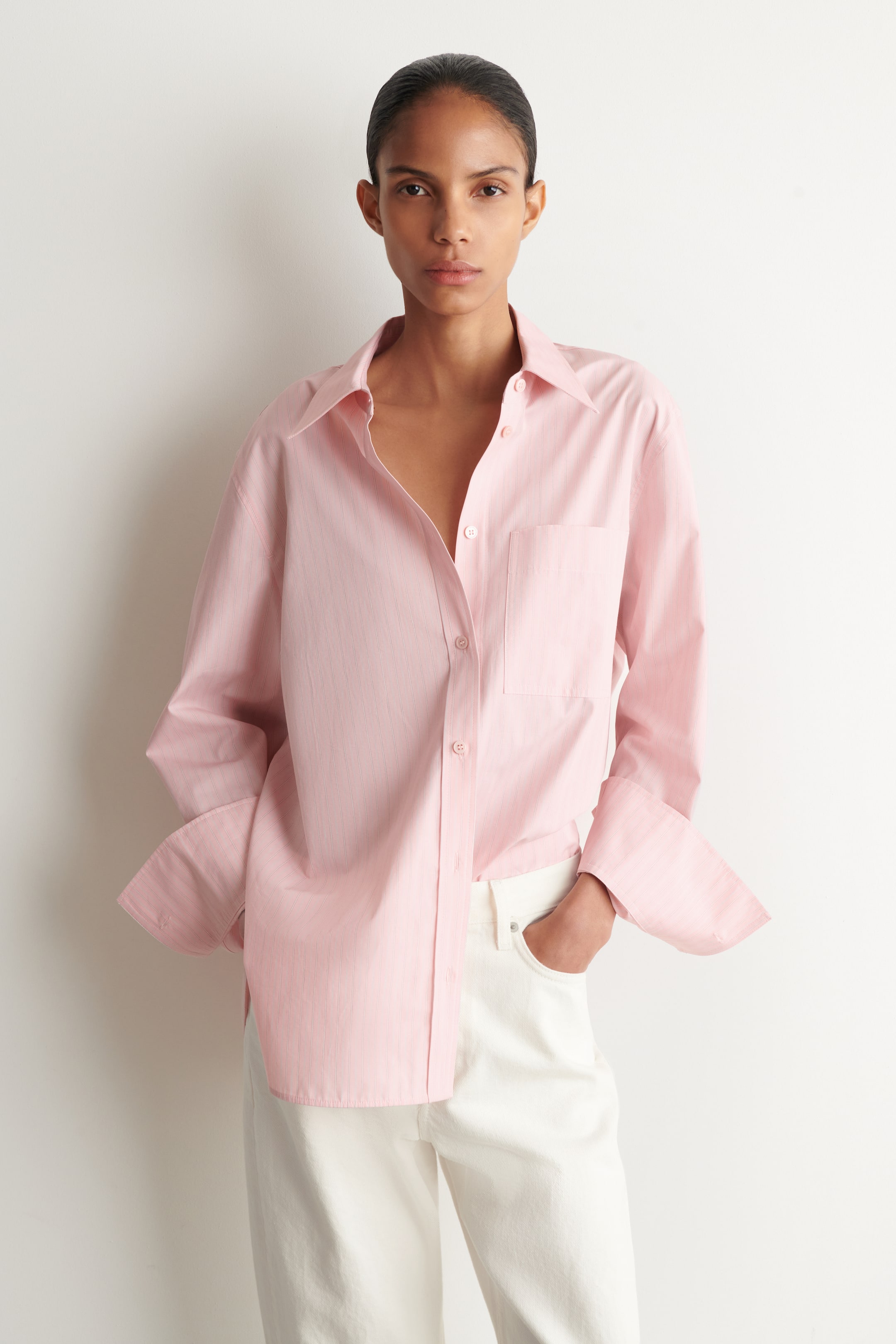 View larger image: A pink pinstriped cotton shirt is displayed as a top, featuring a classic collar, button fastening, and a chest patch pocket. Its long sleeves are styled with wide, turned-back cuffs.