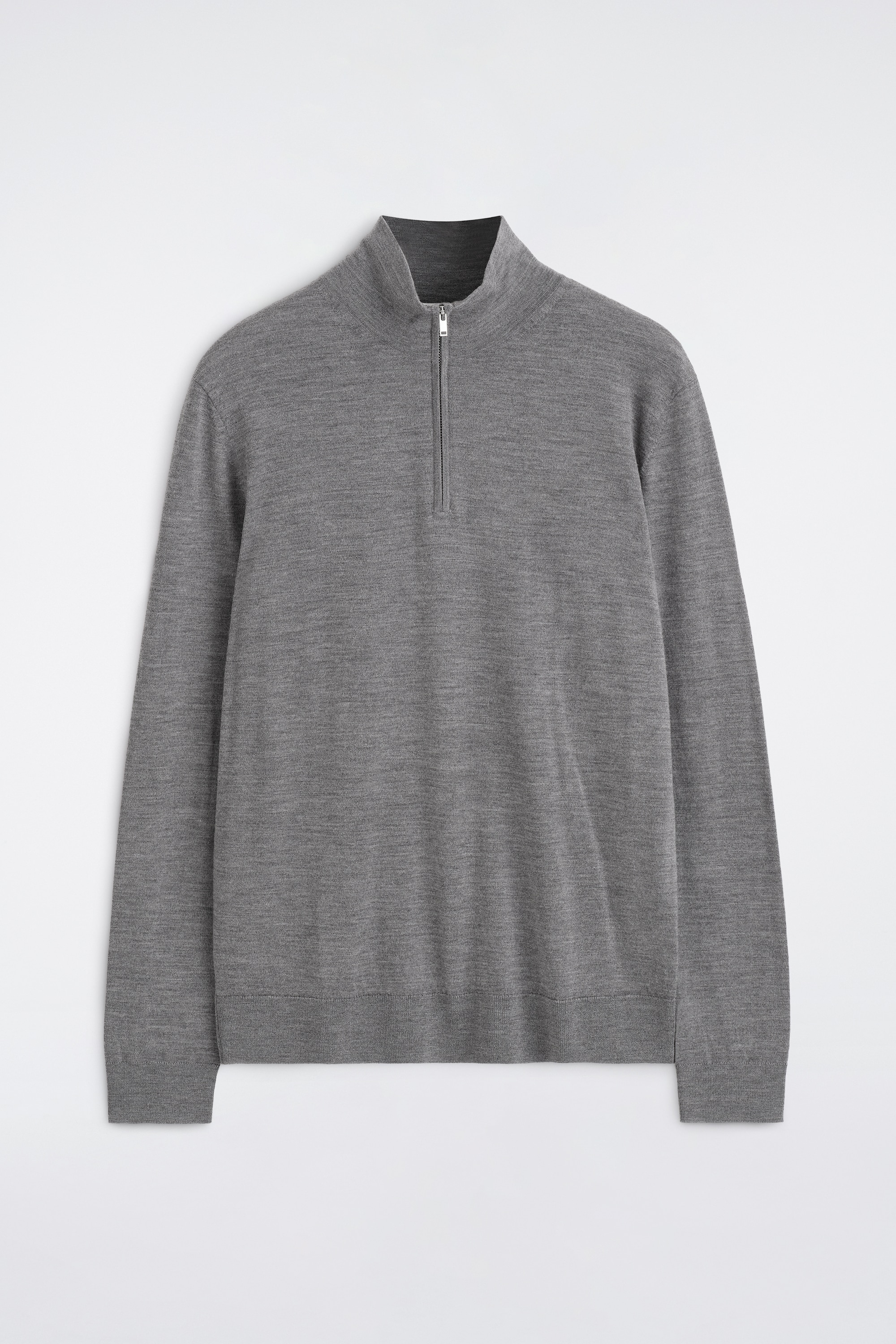 La modella indossa un maglione grigio in lana merino con collo a imbuto e zip, caratterizzato da un colletto con mezza zip argentata parzialmente aperta per mettere in risalto una t-shirt bianca a strati. Il maglione è abbinato a pantaloni neri, con la modella che tiene le mani nelle tasche.