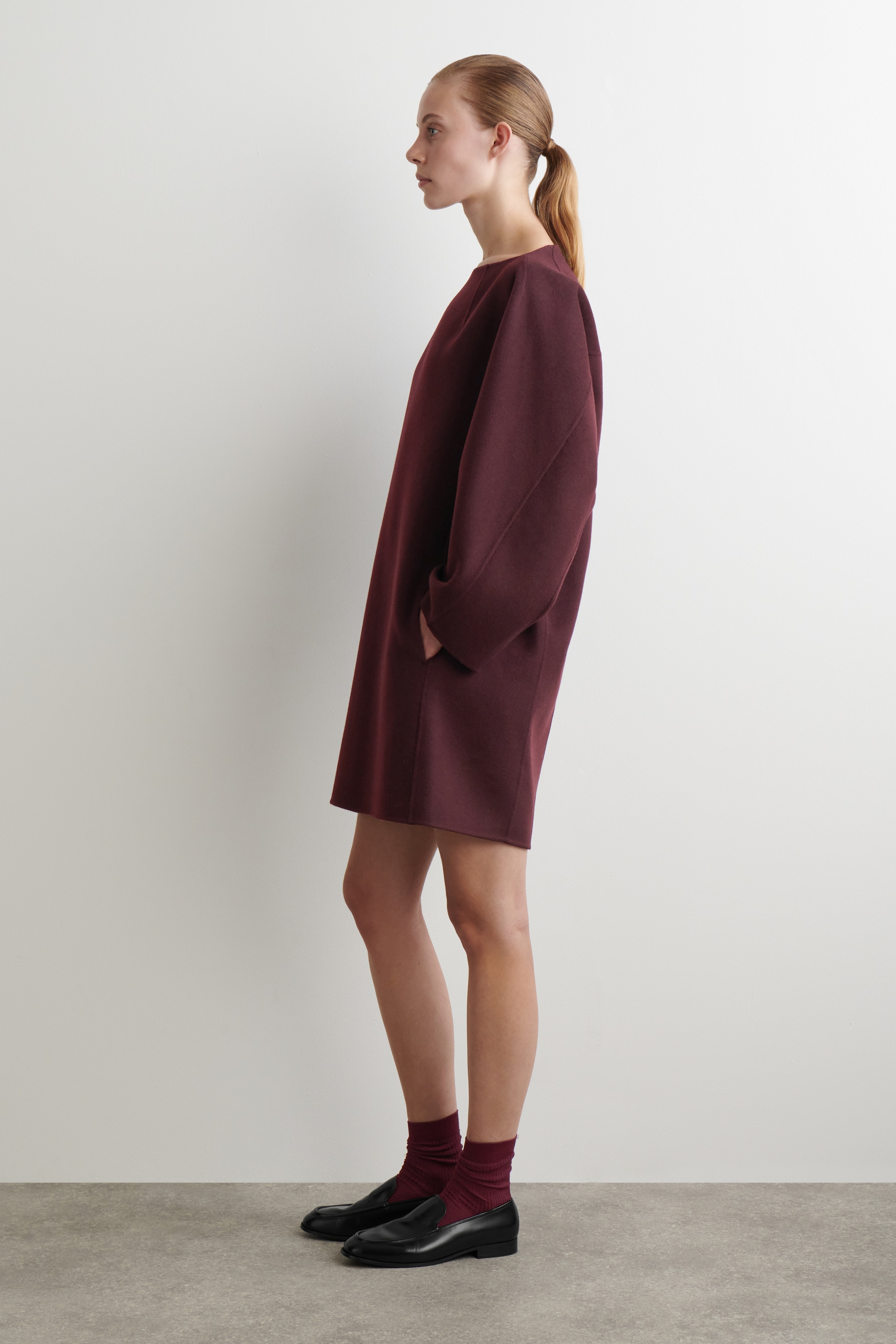 Agrandir l'image: Une robe ample de couleur marron est présentée de profil, mettant en valeur sa coupe décontractée et son design. La robe présente un large décolleté, des manches longues volumineuses avec des épaules tombantes, et se termine au-dessus du genou. Les mains de la personne qui la porte reposent dans des poches latérales, et la robe est assortie de chaussettes marron et de mocassins noirs.