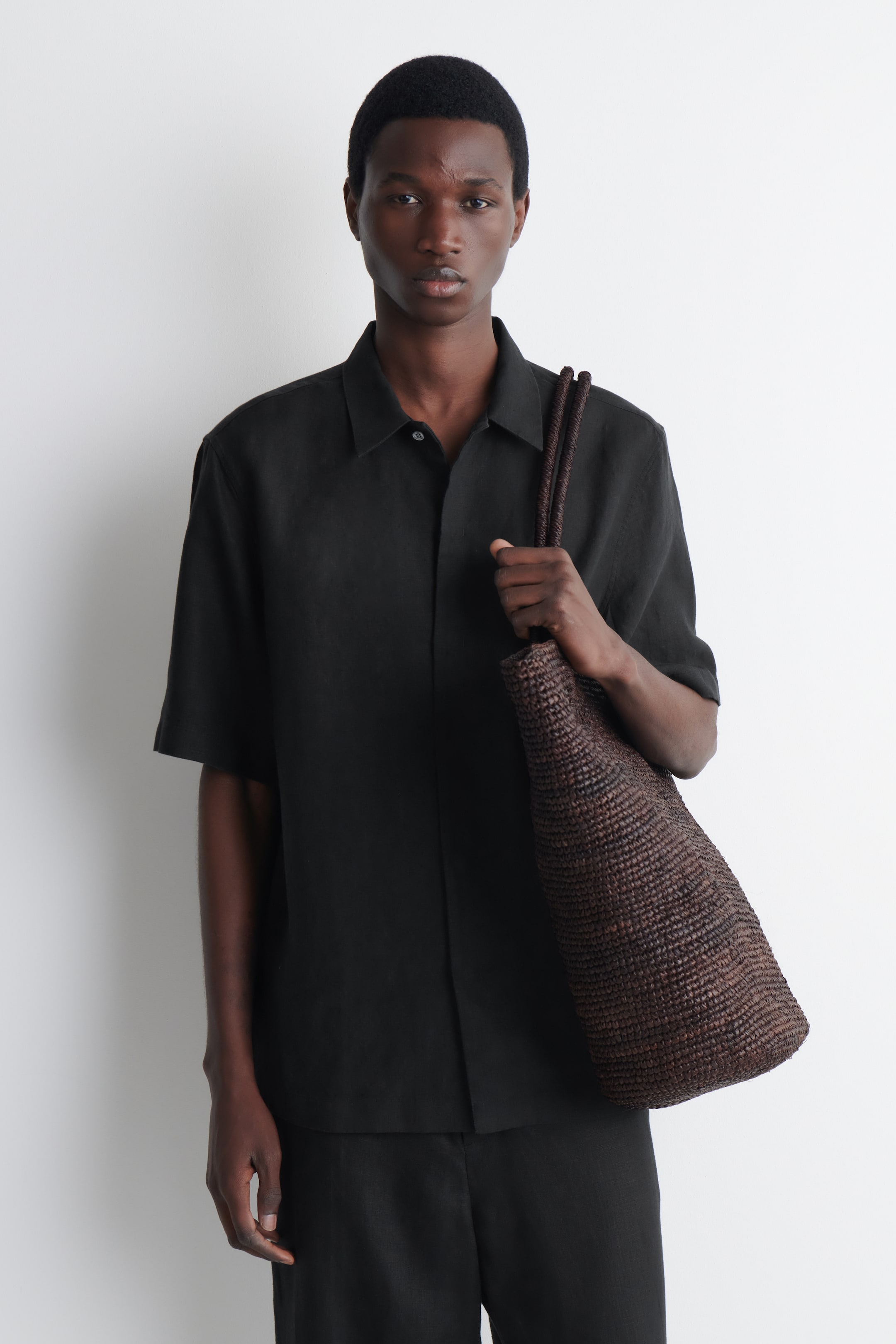 Agrandir l'image: Une chemise noire à manches courtes en lin, dotée d'un col pointu et d'une patte de boutonnage, est portée par le mannequin. Le mannequin porte également un pantalon noir assorti et tient un sac cabas tressé en cuir marron foncé, qui repose contre la chemise.