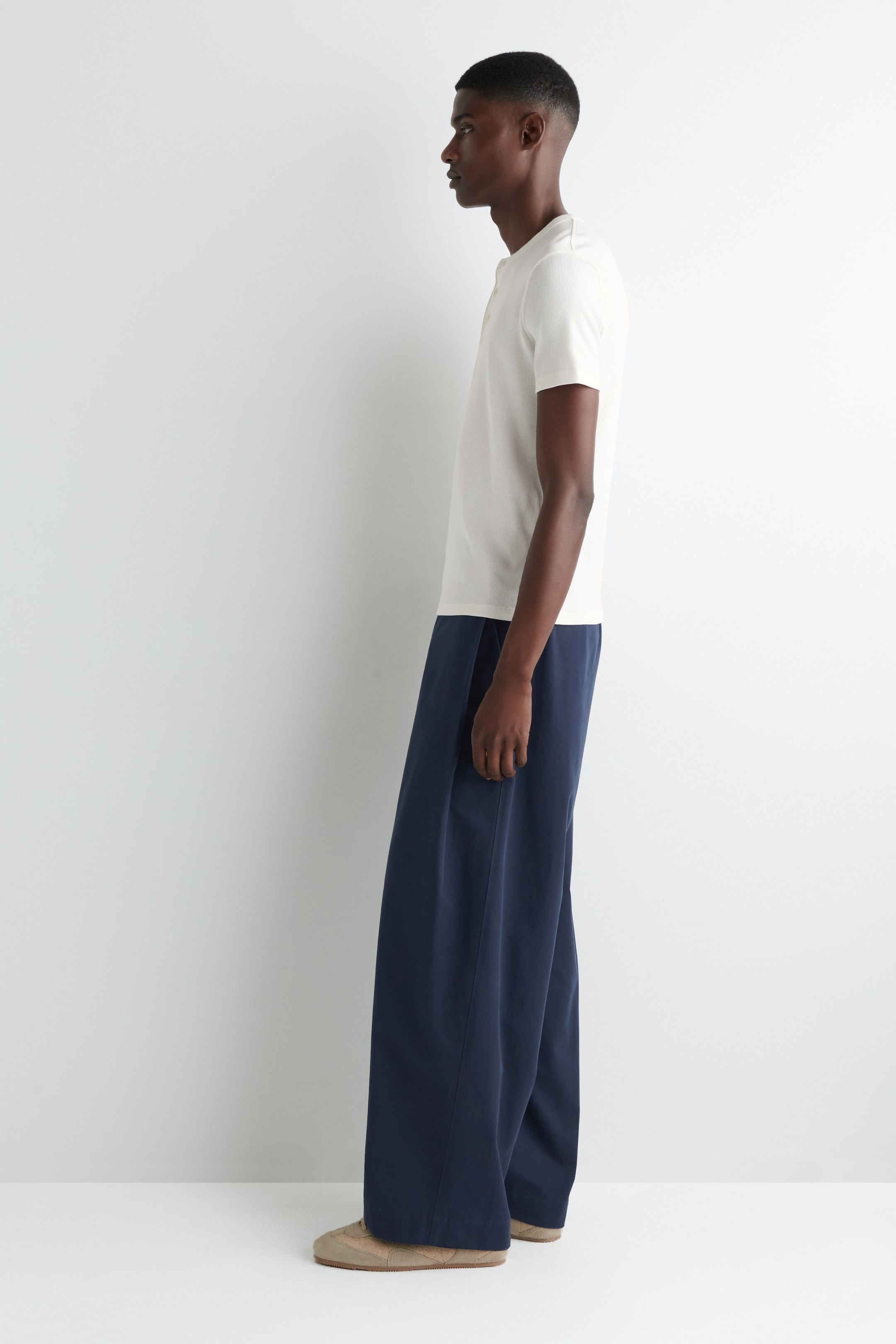 View larger image: A model is wearing a cream short-sleeved Henley shirt and navy wide-leg pants, presented in a side profile view. The shirt is featuring a buttoned placket and a regular fit, while the pants are showcasing a loose, flowing silhouette.