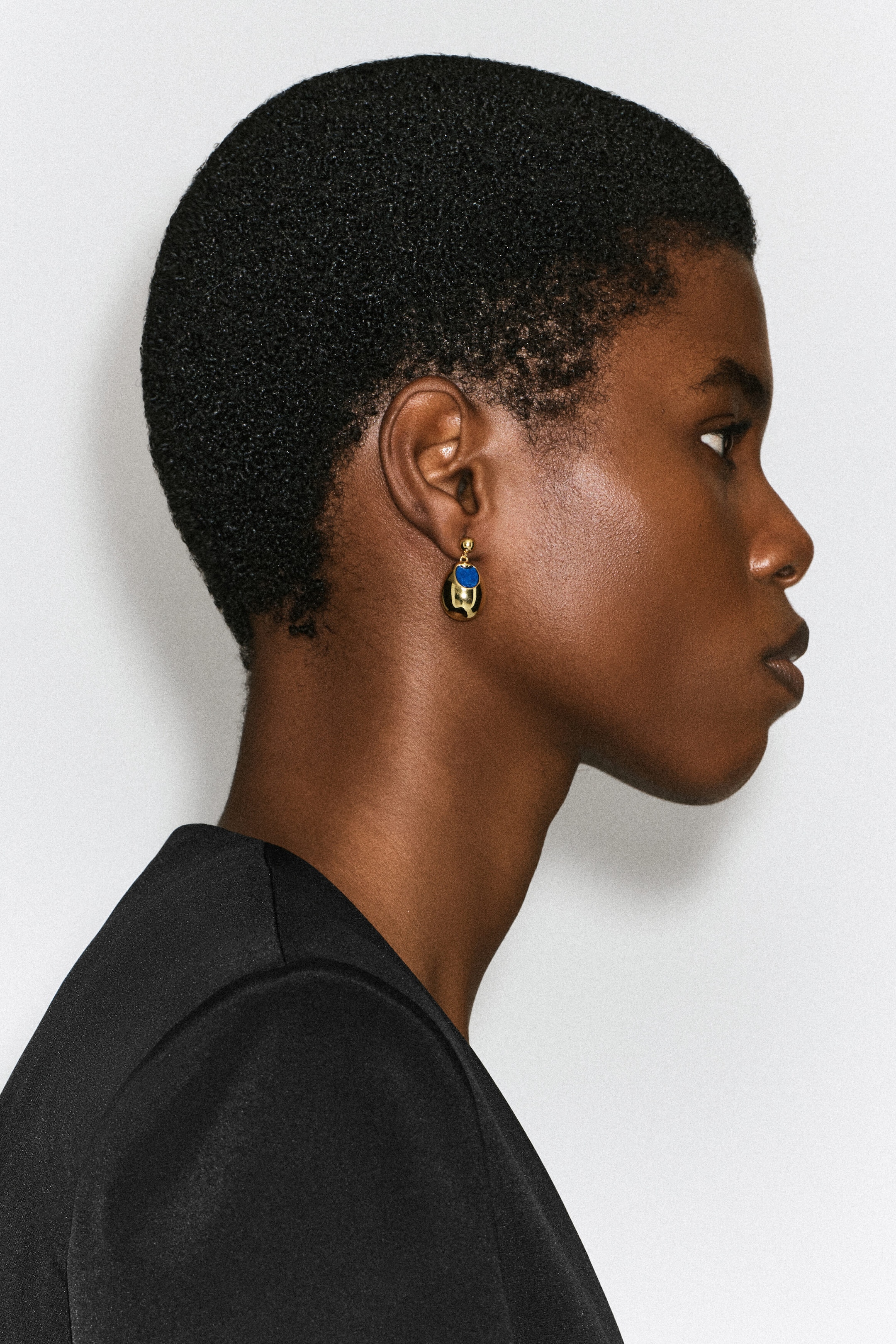 Le mannequin porte une boucle d’oreille pendante détaillée de pierres, en vue de profil. La boucle d’oreille présente un petit clou doré et un pendentif doré arrondi en forme de goutte, avec une pierre bleue incrustée.
