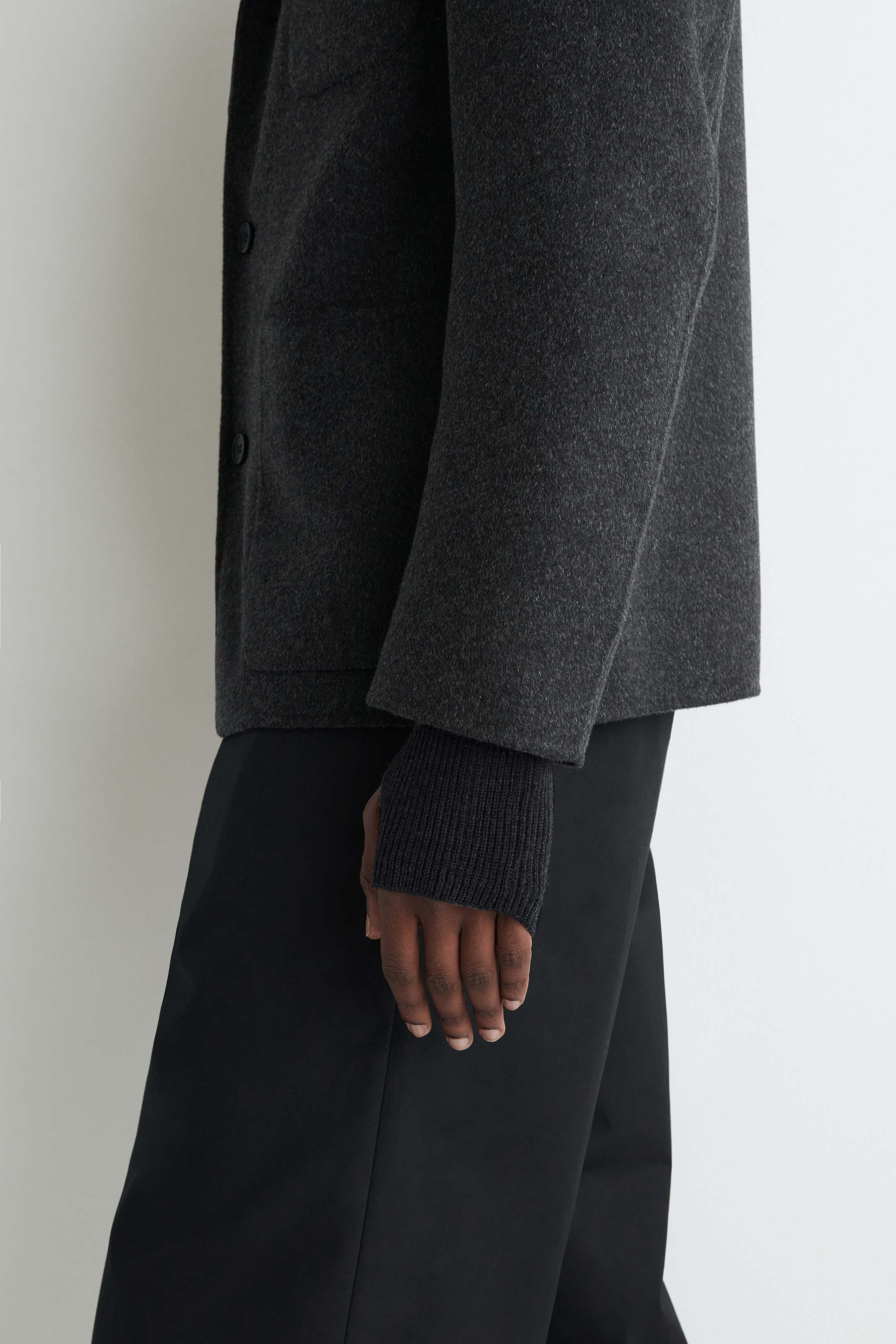 Agrandir l'image: Une veste gris foncé est vue de côté, avec un tissu texturé et des boutons sombres visibles sur sa patte de boutonnage avant. Ses manches larges se terminent au-dessus du poignet, révélant les poignets côtelés d'un haut à manches longues gris foncé en dessous. Un pantalon noir à jambes larges est également porté.