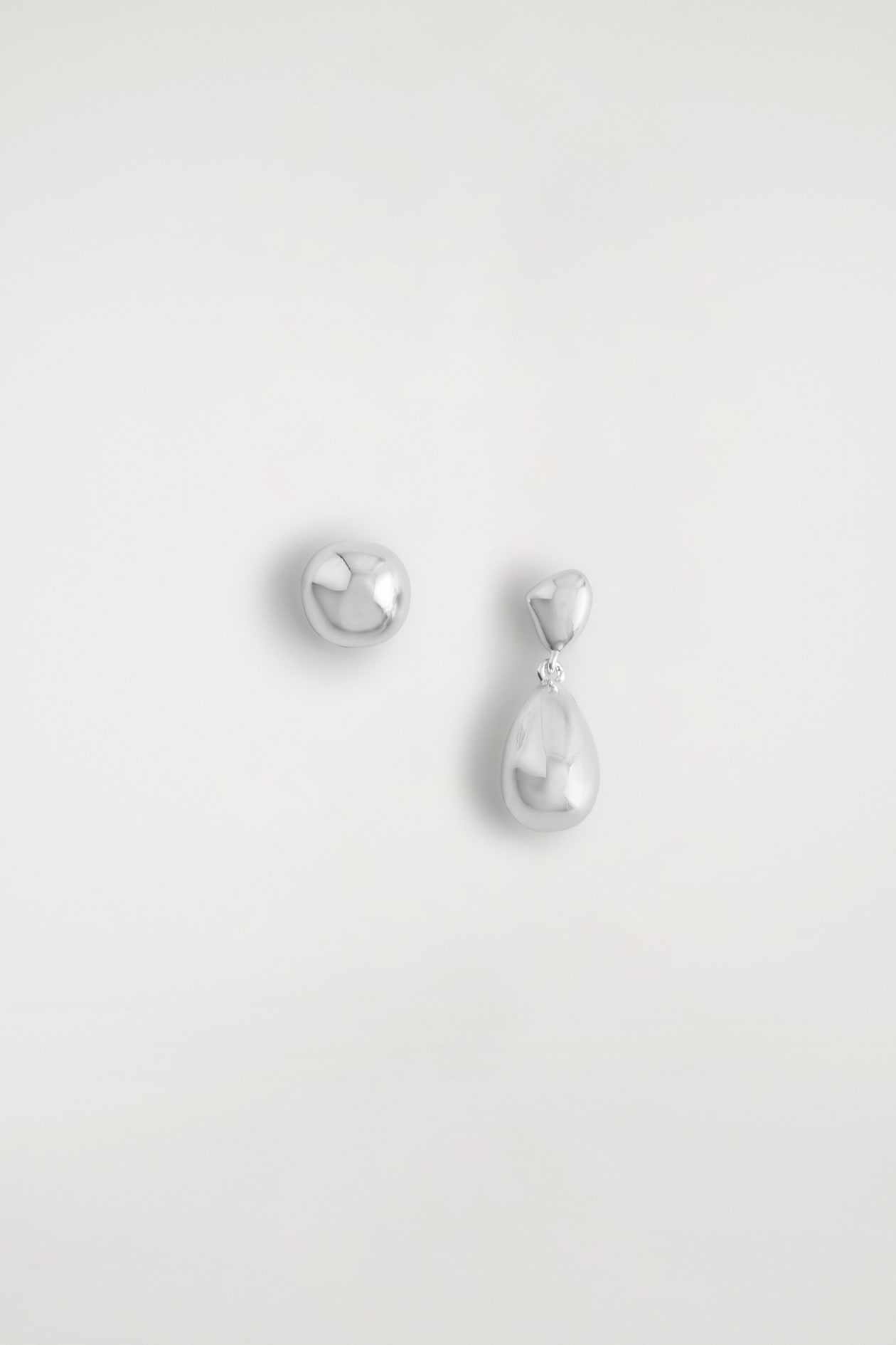 Mismatched drop earrings are shown from the front, featuring a highly polished silver-tone finish. One earring is shown as a single, rounded organic-shaped stud, while the other features a smaller stud suspending a smooth, teardrop-shaped pendant.
