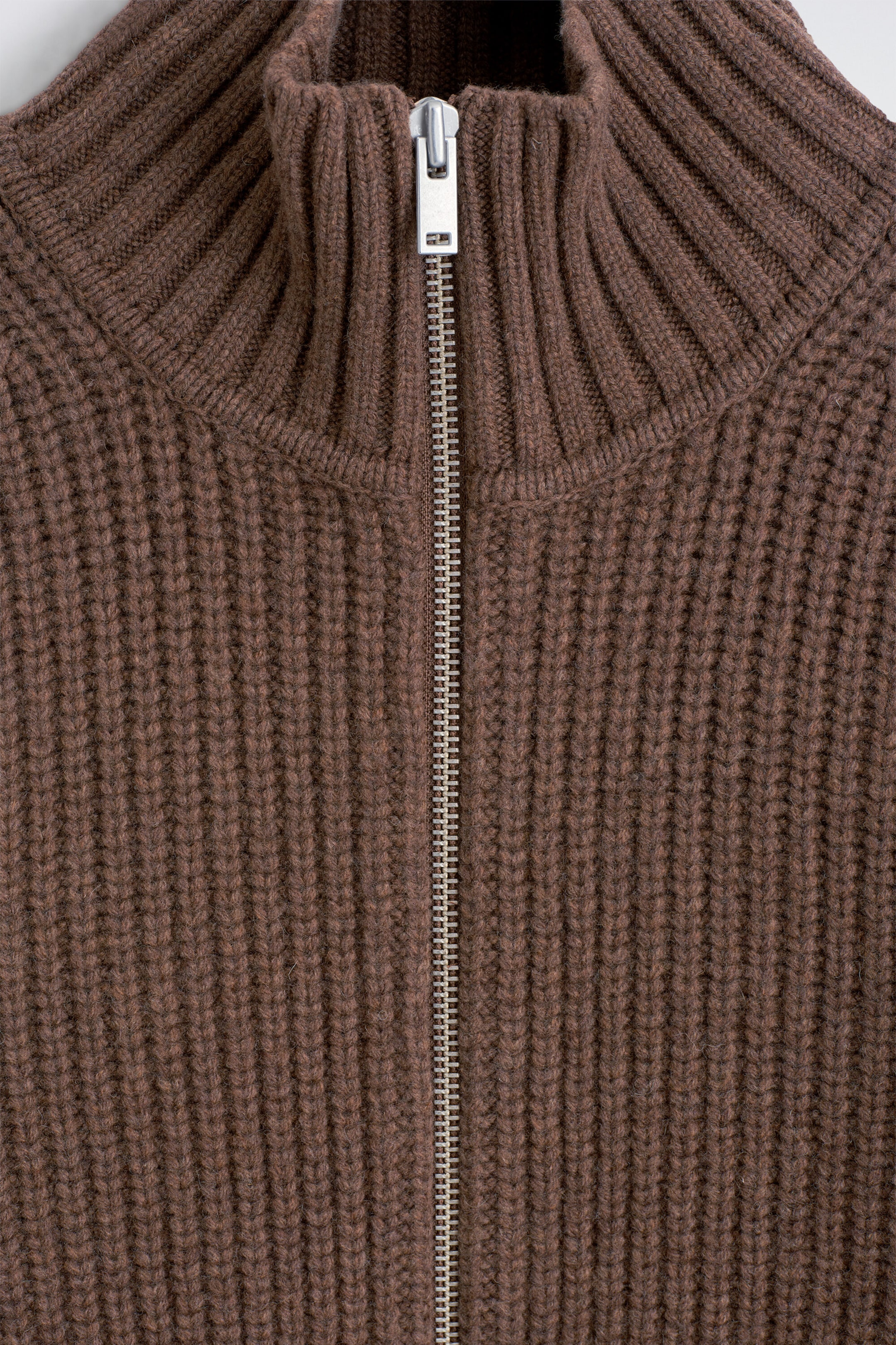 A brown ribbed knit sweater features a high mock neck collar with vertical ribbing and a central silver-toned metal zipper. The garment displays a textured, chunky ribbed knit pattern throughout the body. A brown ribbed knit sweater features a high mock neck collar with vertical ribbing and a central silver-toned metal zipper. The garment displays a textured, chunky ribbed knit pattern throughout the body.